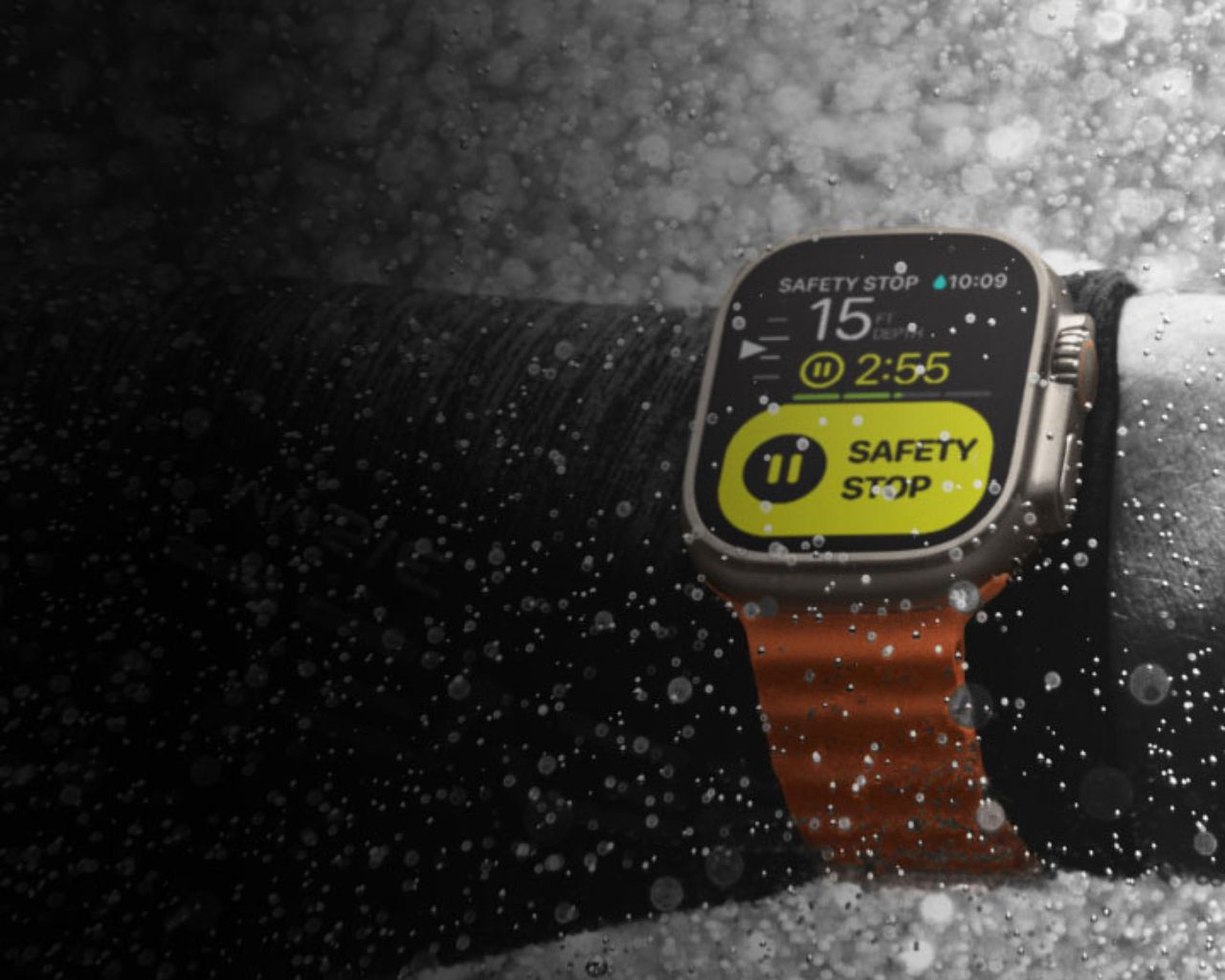 Inteligentné hodinky na zápästie s jasne oranžovým remienkom, ktoré zobrazujú časovač „Bezpečnostného zastavenia“ na tmavom, vodou postriekanom pozadí.