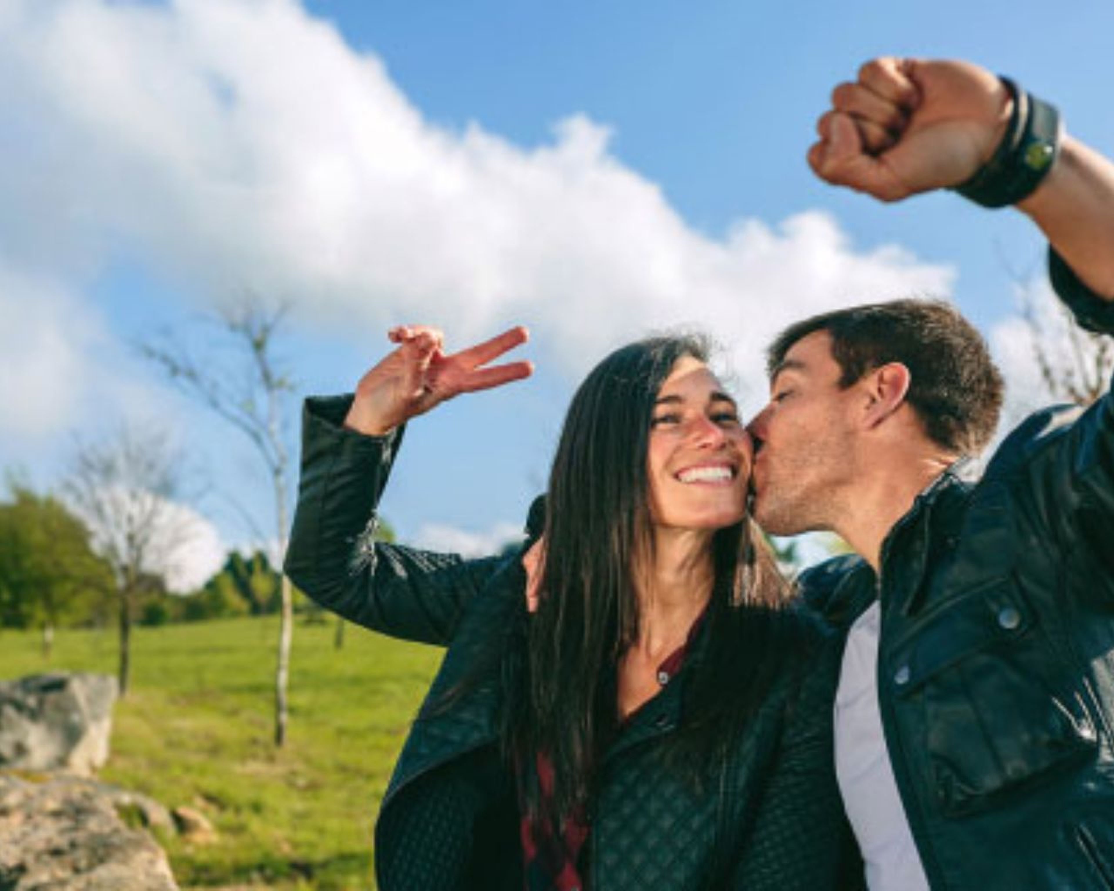 Pár pózuje vonku; muž pobozká ženu na líce, zatiaľ čo sa ona usmieva a ukazuje znamenie mieru. Majú na sebe bundy a za nimi je trávnaté pole.