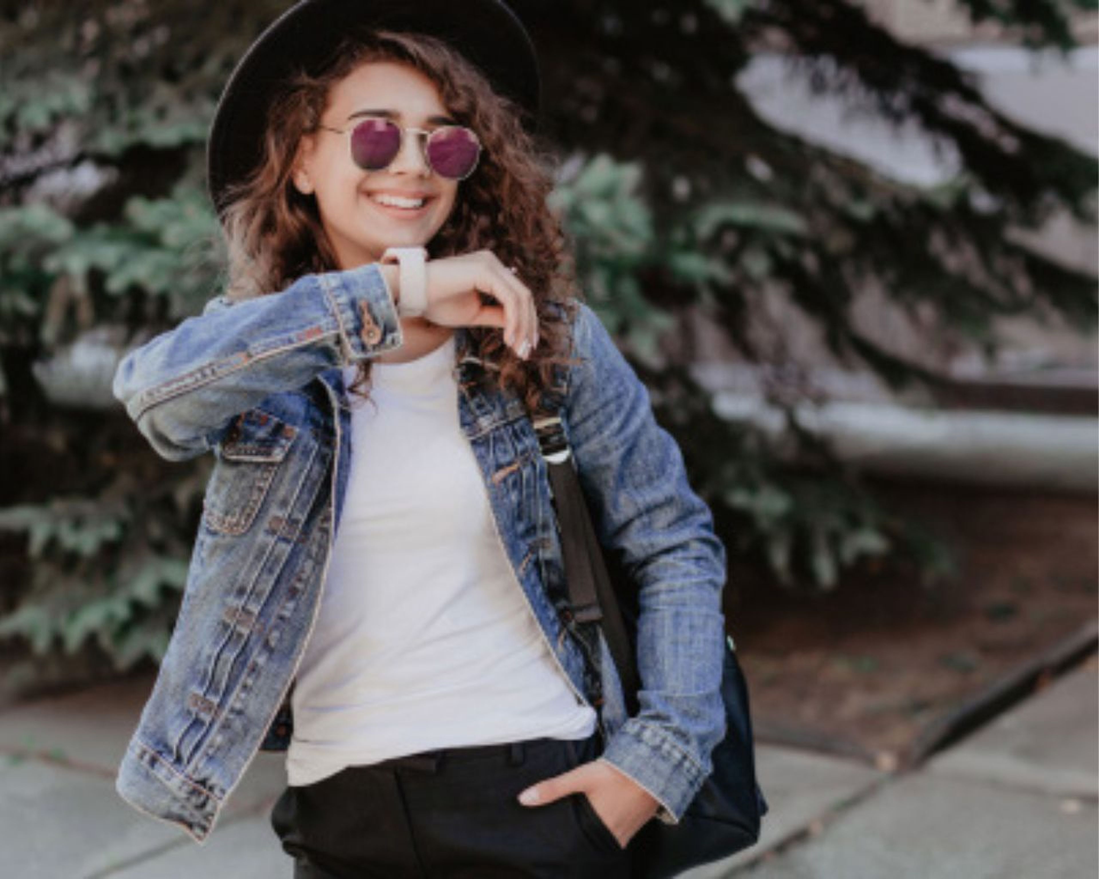 Usmievavá žena v džínsovej bunde, bielej košeli a slnečných okuliaroch, s čiernym klobúkom, stojí vonku s rukou vo vrecku a taškou cez plece.