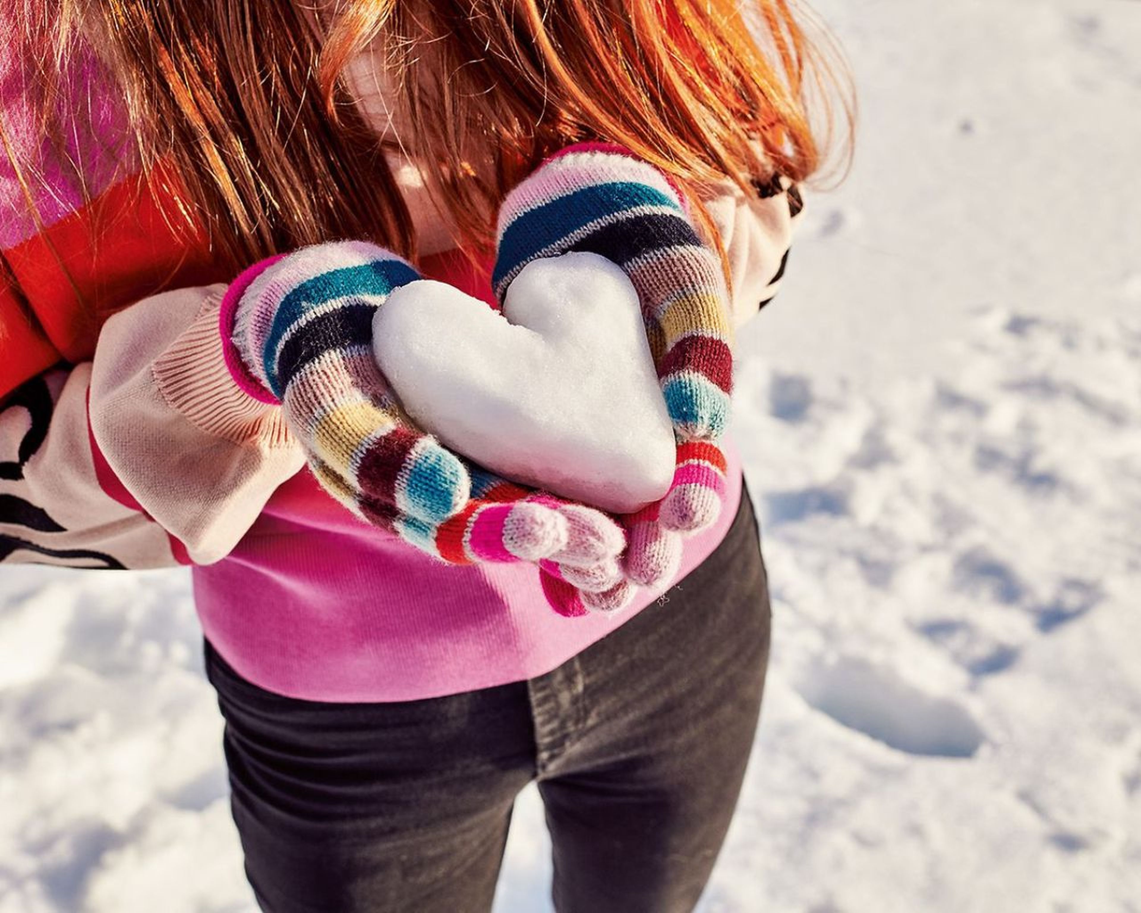 Osoba vo farebných rukaviciach drží snehovú guľu v tvare srdca a stojí na zasneženej zemi.