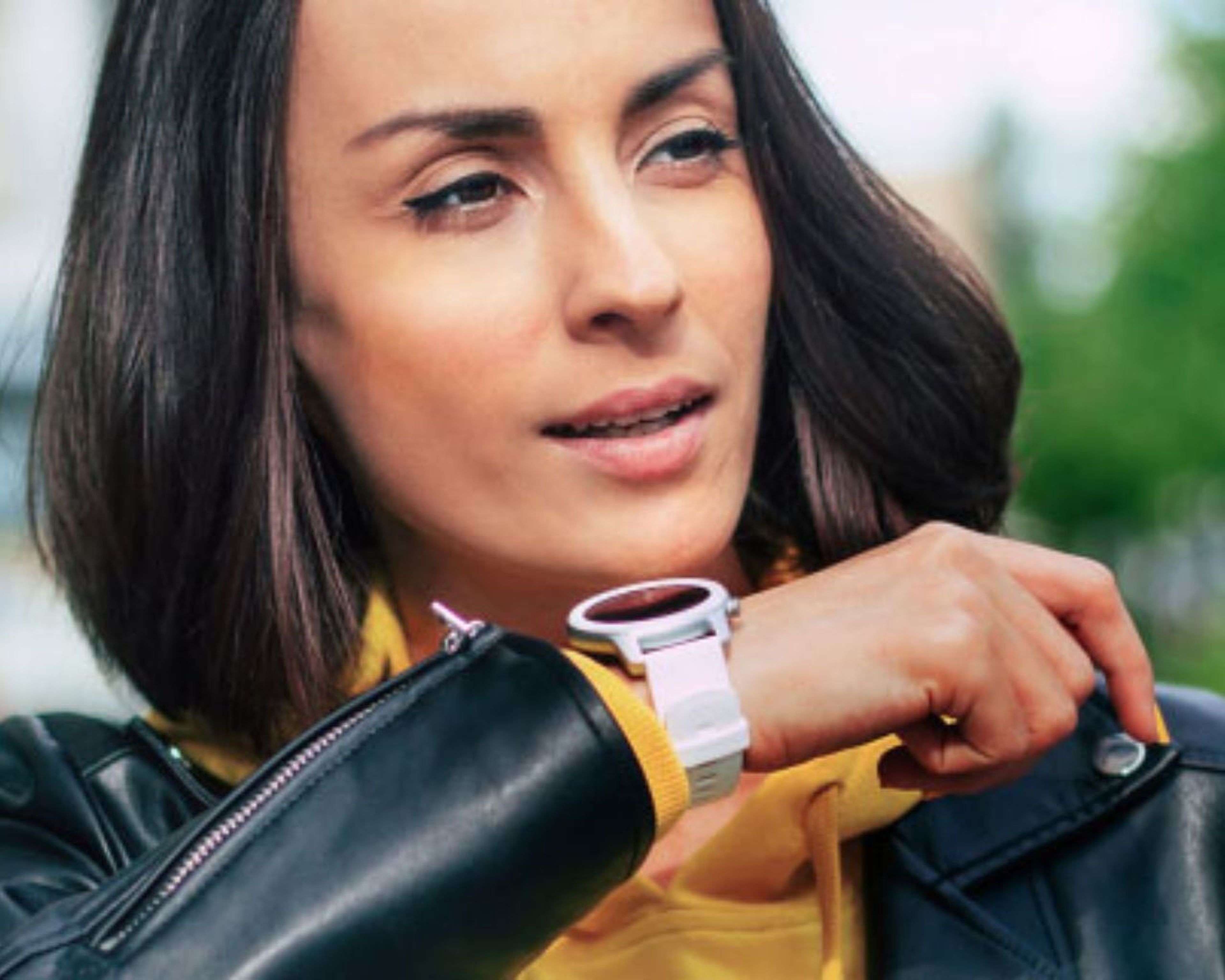Osoba vonku v čiernej koženej bunde a žltej mikine s kapucňou, ktorá sa sústredene pozerá na inteligentné hodinky.