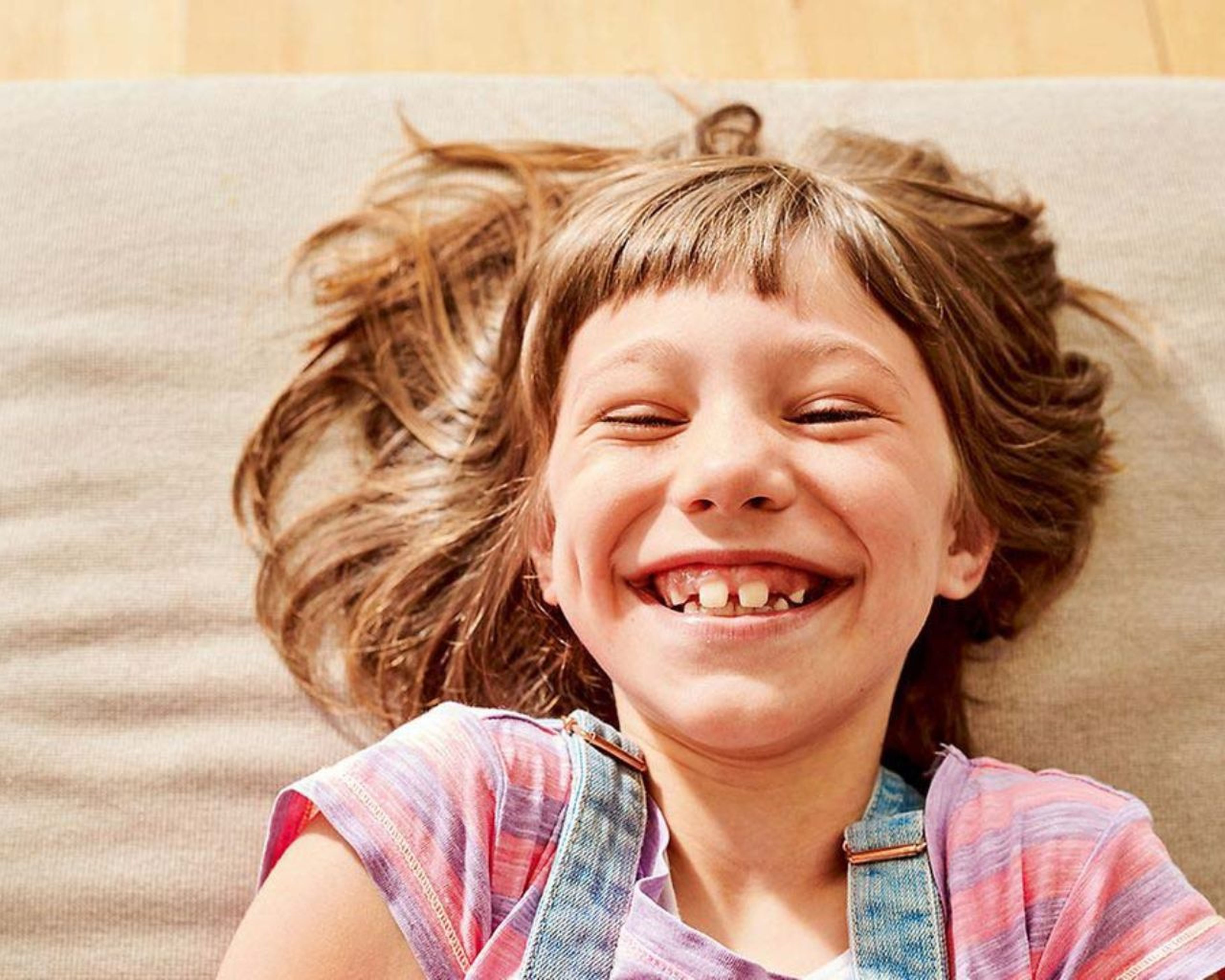 Usmievavé dieťa s hnedými vlasmi leží na svetlom vankúši, má na sebe ružové tričko a džínsový overal.