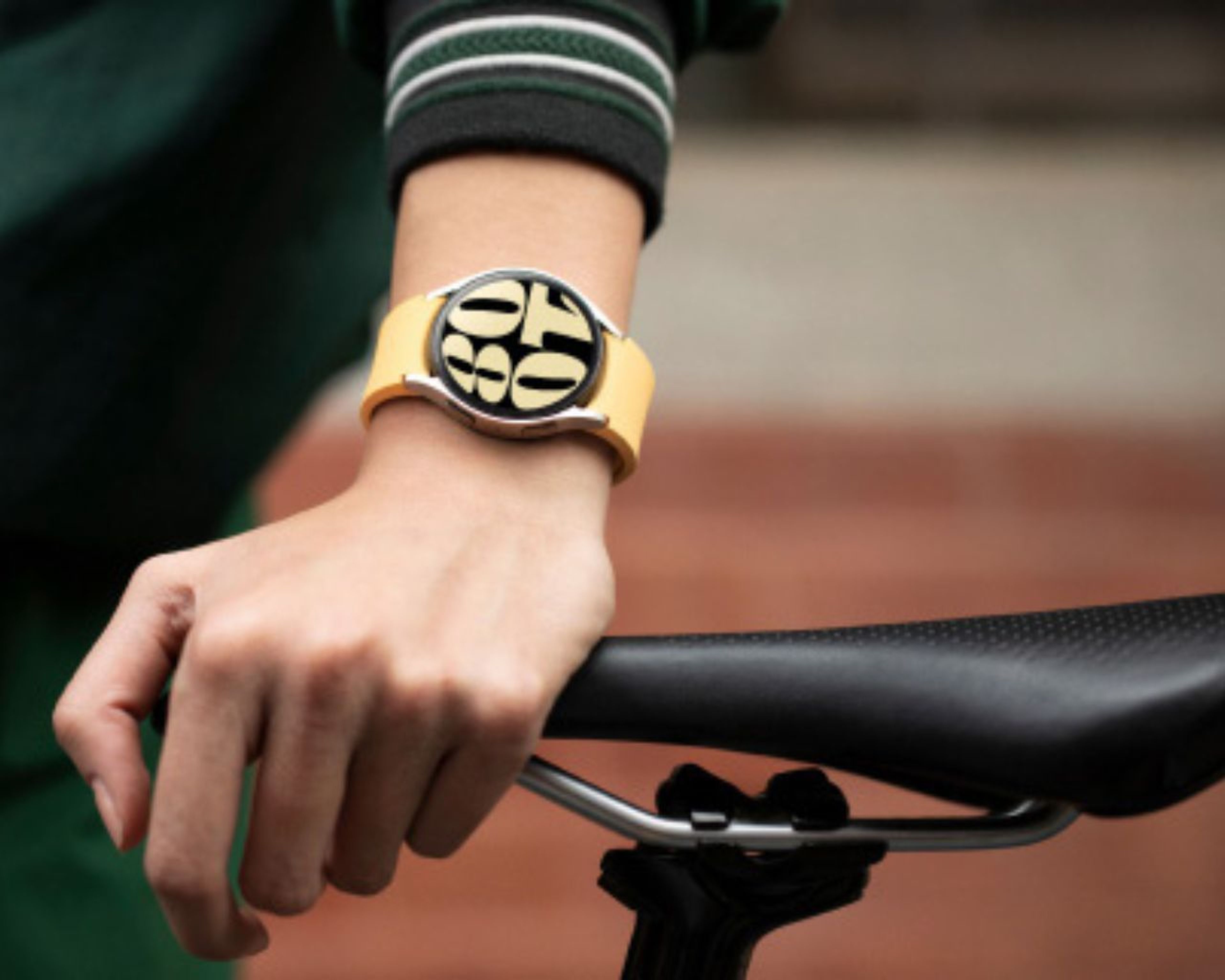 Osoba so žltými hodinkami s výraznými číslicami, rukou položenou na sedadle bicykla a oblečenou zelenou bundou.