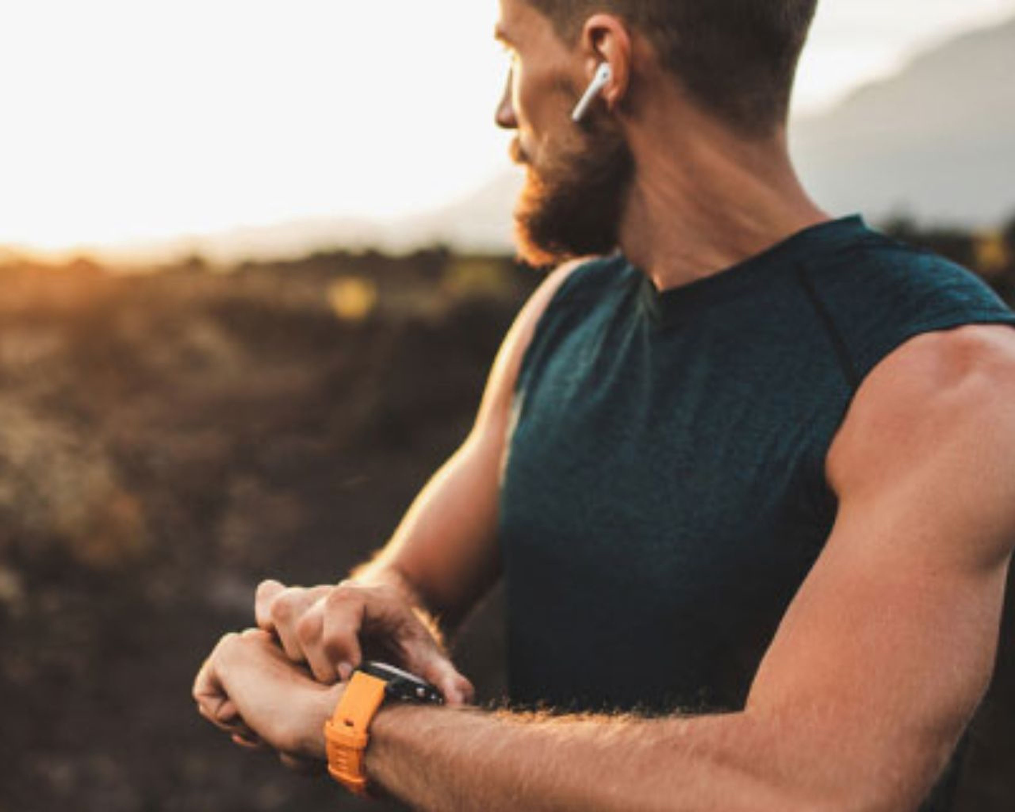 Muž s bradou, slúchadlami do uší a tričkom bez rukávov si kontroluje inteligentné hodinky vonku pri západe slnka, v pozadí hory.