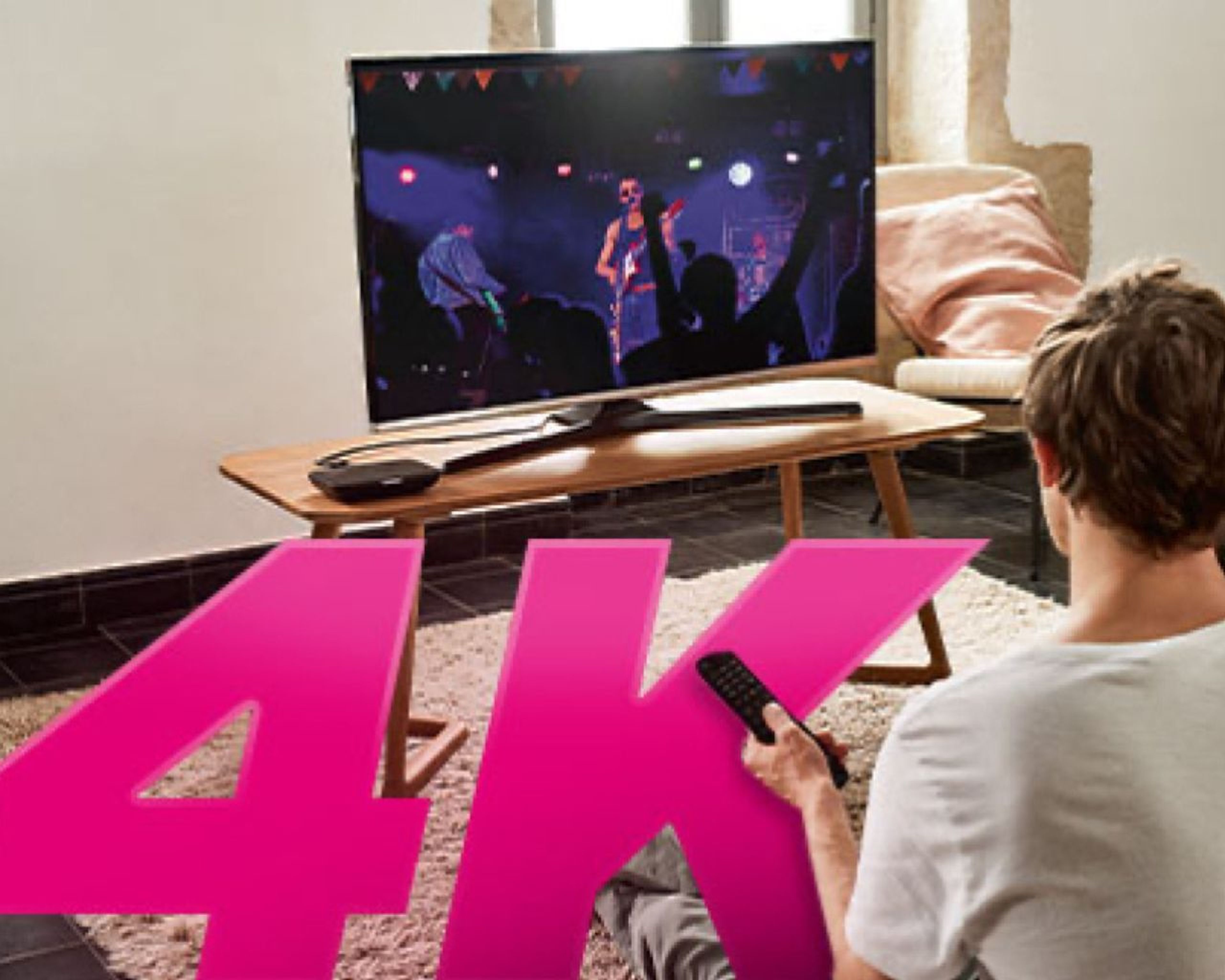 Osoba pozerá koncert na 4K televízore a drží v ruke diaľkové ovládanie. Na drevenom stole pred televízorom je soundbar.