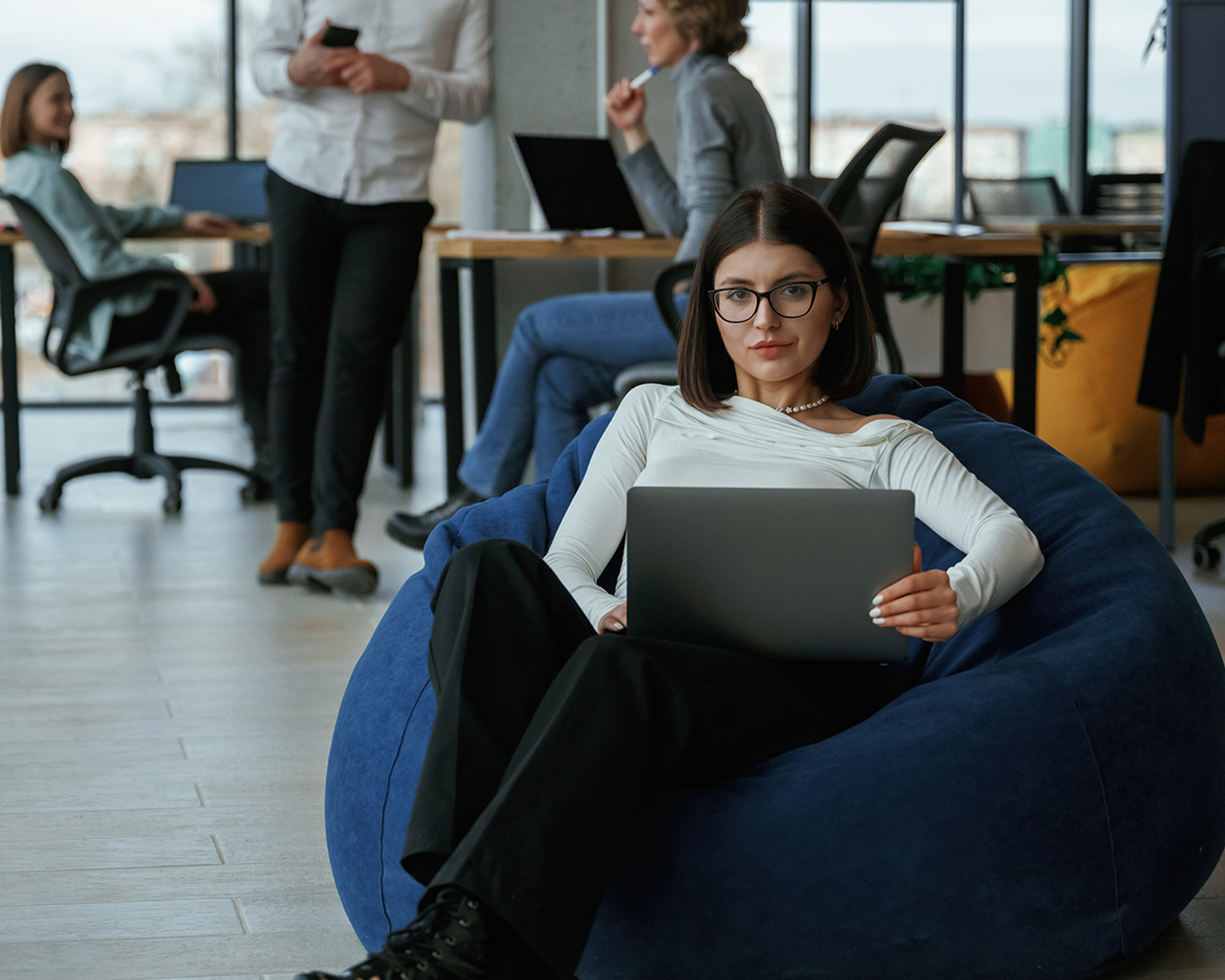 Žena s okuliarmi sediaca s notebookom na kolenách v modernej kancelárii. V pozadí je vidno ďalších kolegov sediacich pri stole.