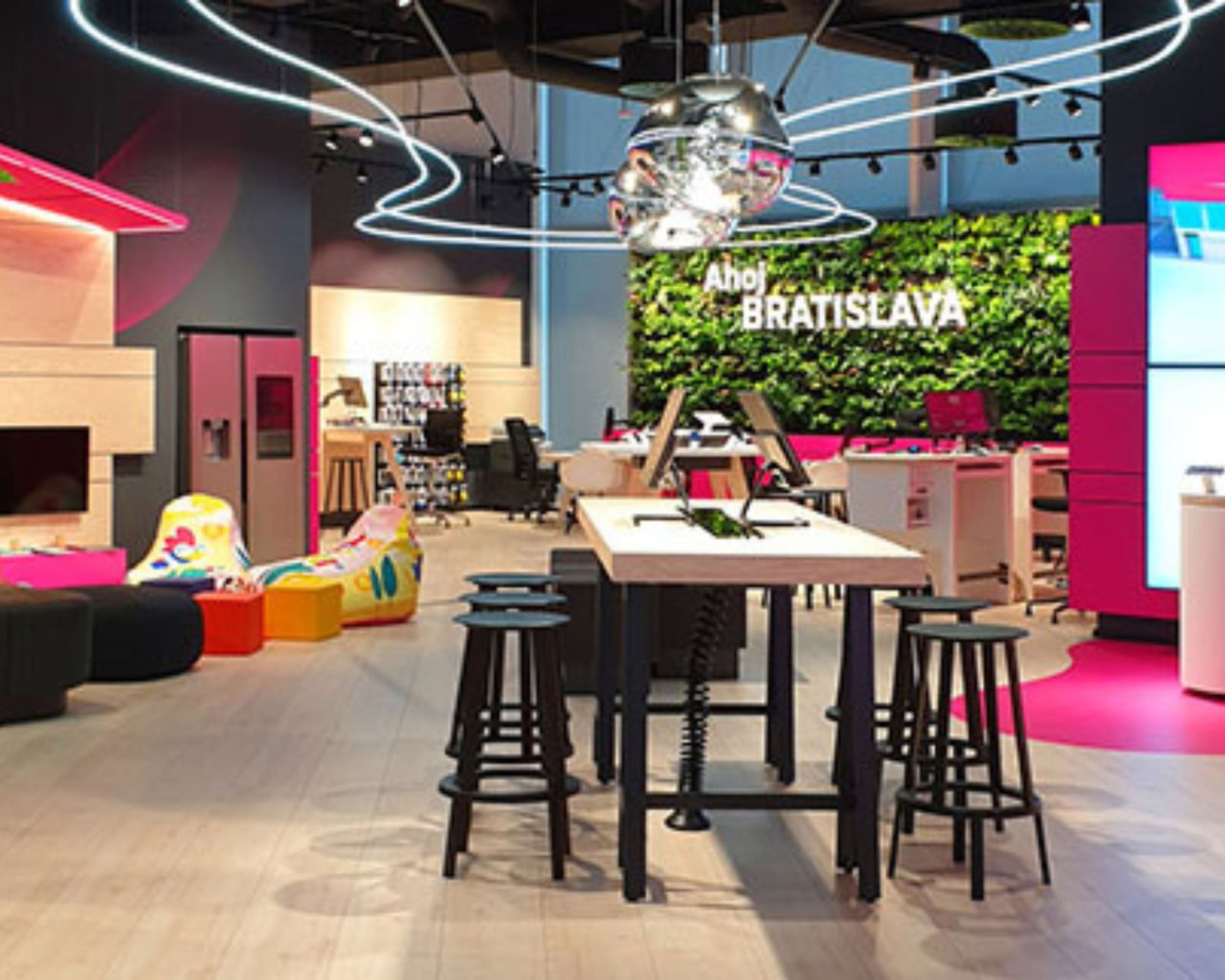 Moderný interiér predajne Telekom s centrálnym stolom, stoličkami, neónovými svetlami a zelenou stenou s nápisom „Ahoj Bratislava“. Svetlá a farebná výzdoba.