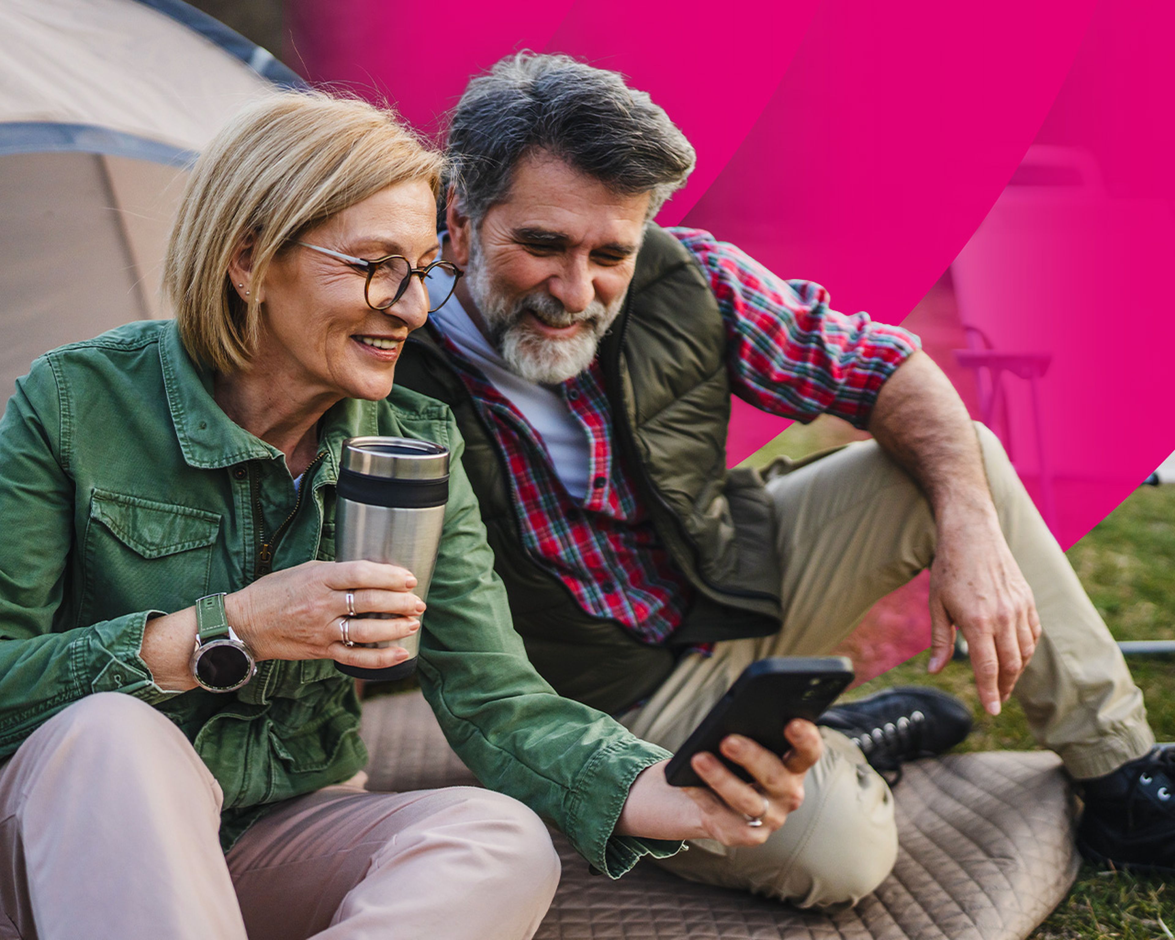 Starší pár sedí na deke vonku pri stane, usmieva sa a pozerá sa do smartfónu. Žena drží cestovný hrnček.