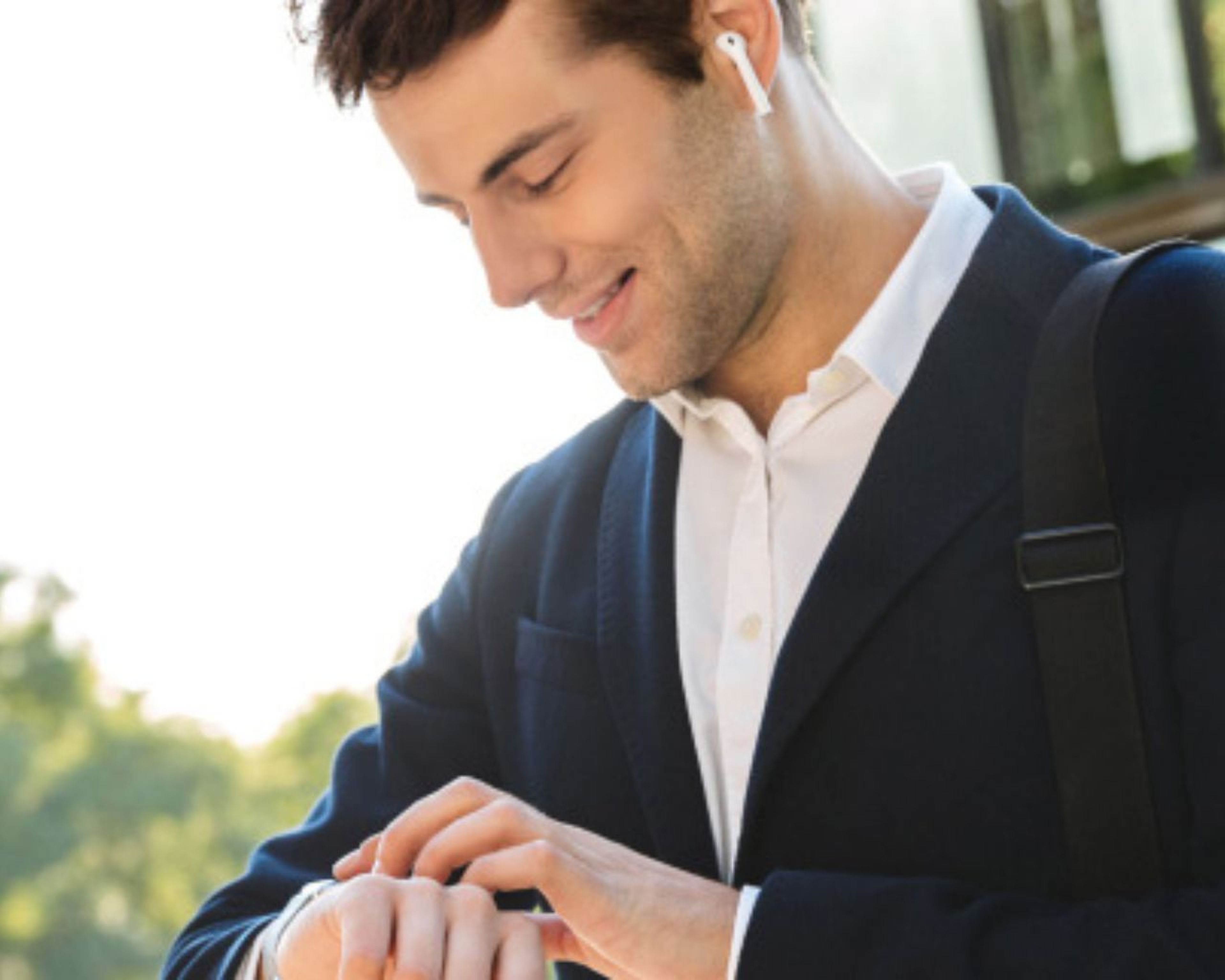 Muž v obleku, ktorý sa pozerá na inteligentné hodinky, má na sebe bezdrôtové slúchadlá a tašku cez rameno, stojí vonku so zeleňou v pozadí.