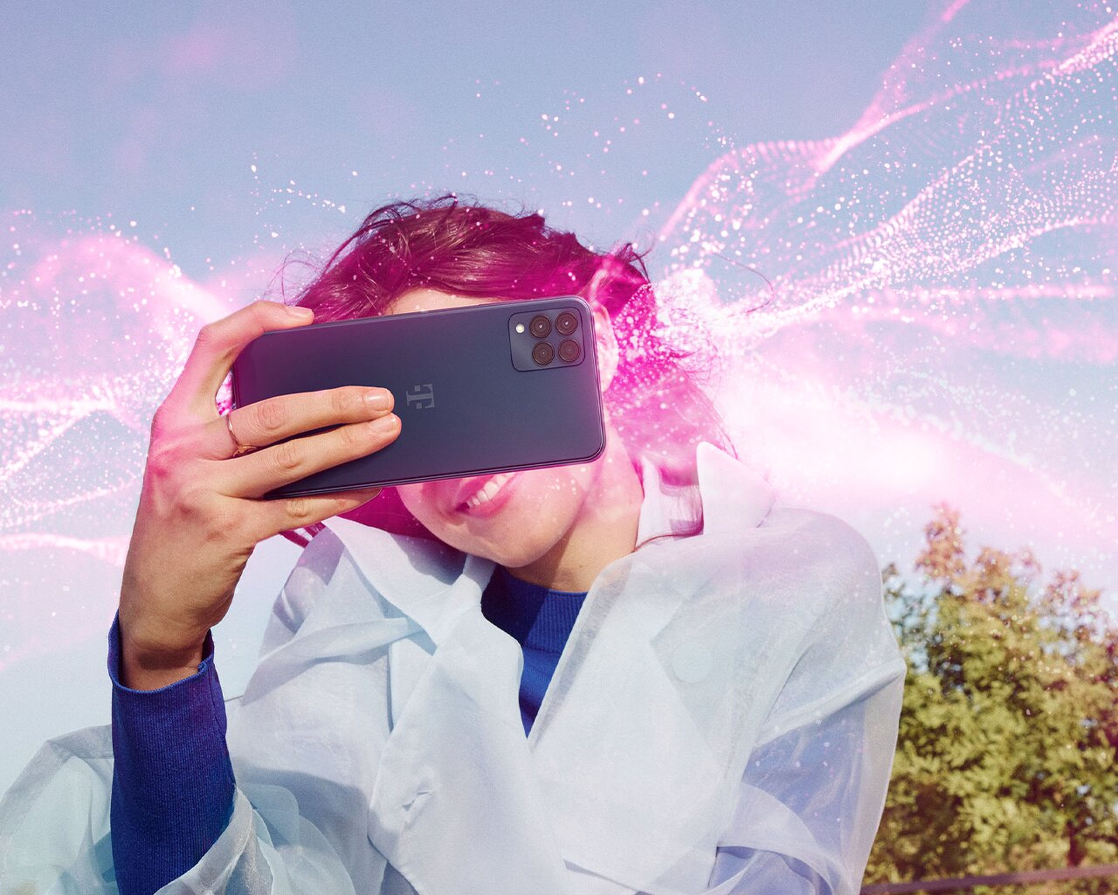 Osoba si robí selfie so smartfónom, zatiaľ čo zo zariadenia vychádzajú žiarivé ružové digitálne efekty na pozadí jasnej oblohy a stromov.