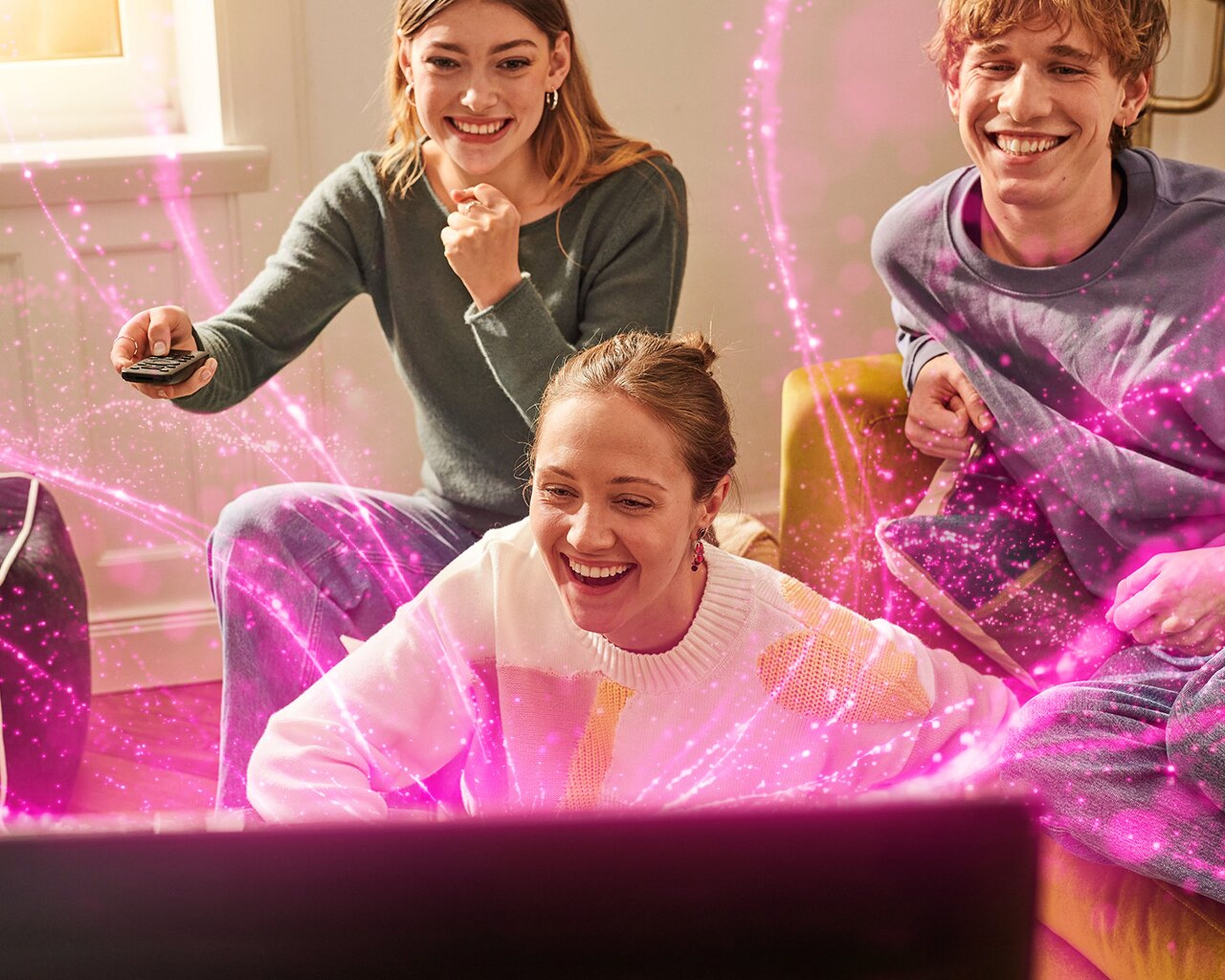 Traja ľudia sediaci na gauči, radostne pozerajú televíziu, obklopení ružovými žiariacimi svetelnými efektmi.