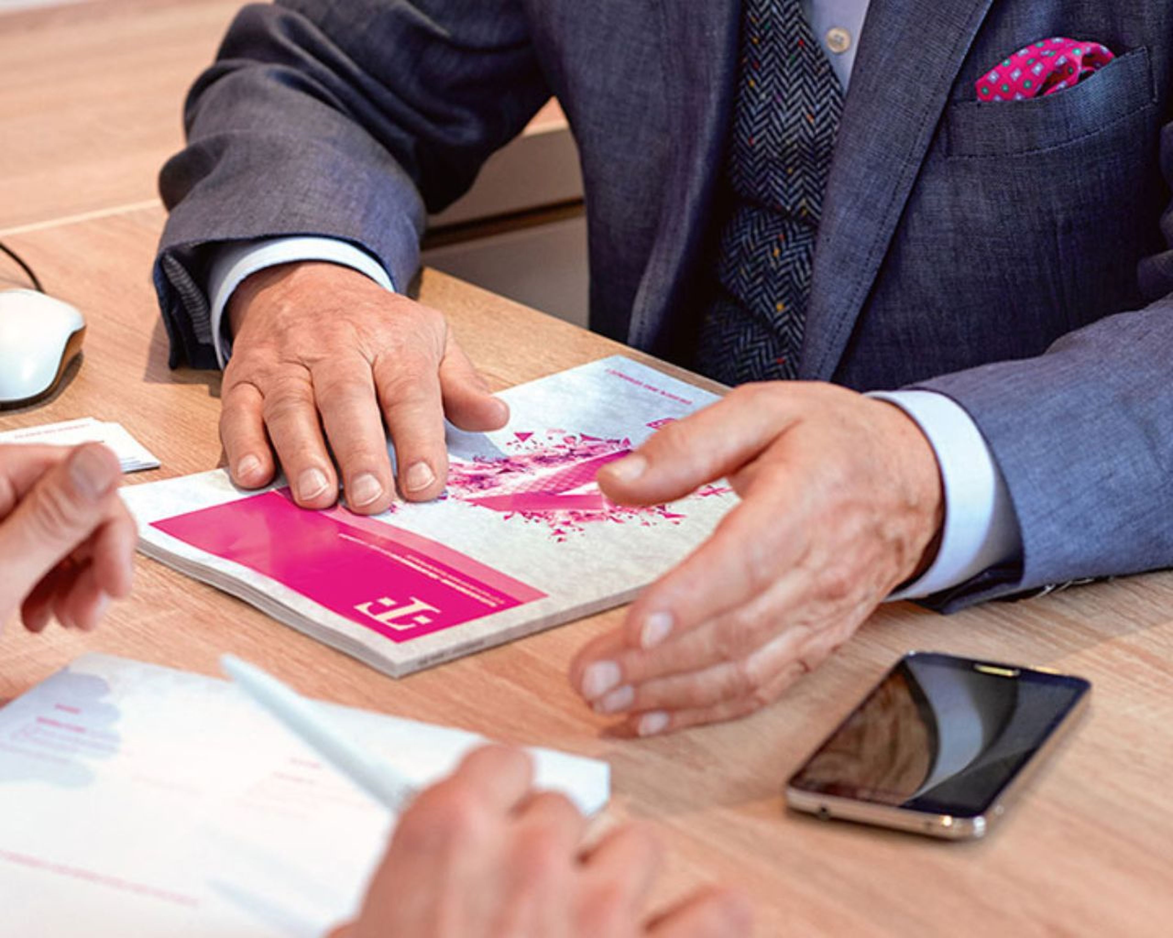 Dvaja ľudia na obchodnom stretnutí, jeden drží pero, druhý má na stole brožúru, vedľa seba smartfón a počítačovú myš.