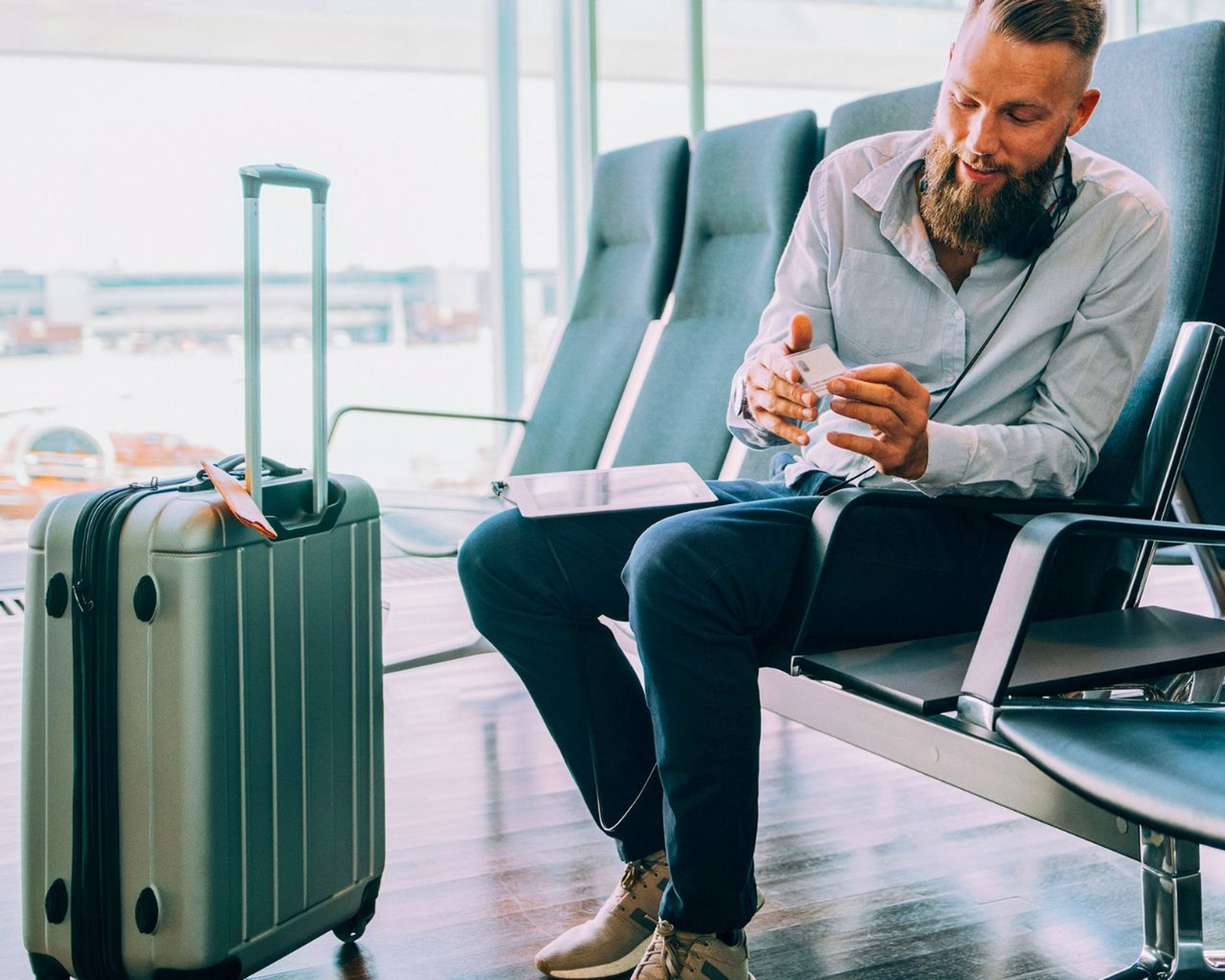 Muž s bradou sedí v letiskovej hale a kontroluje si telefón. Vedľa neho leží kufor a na kolenách má palubný lístok.
