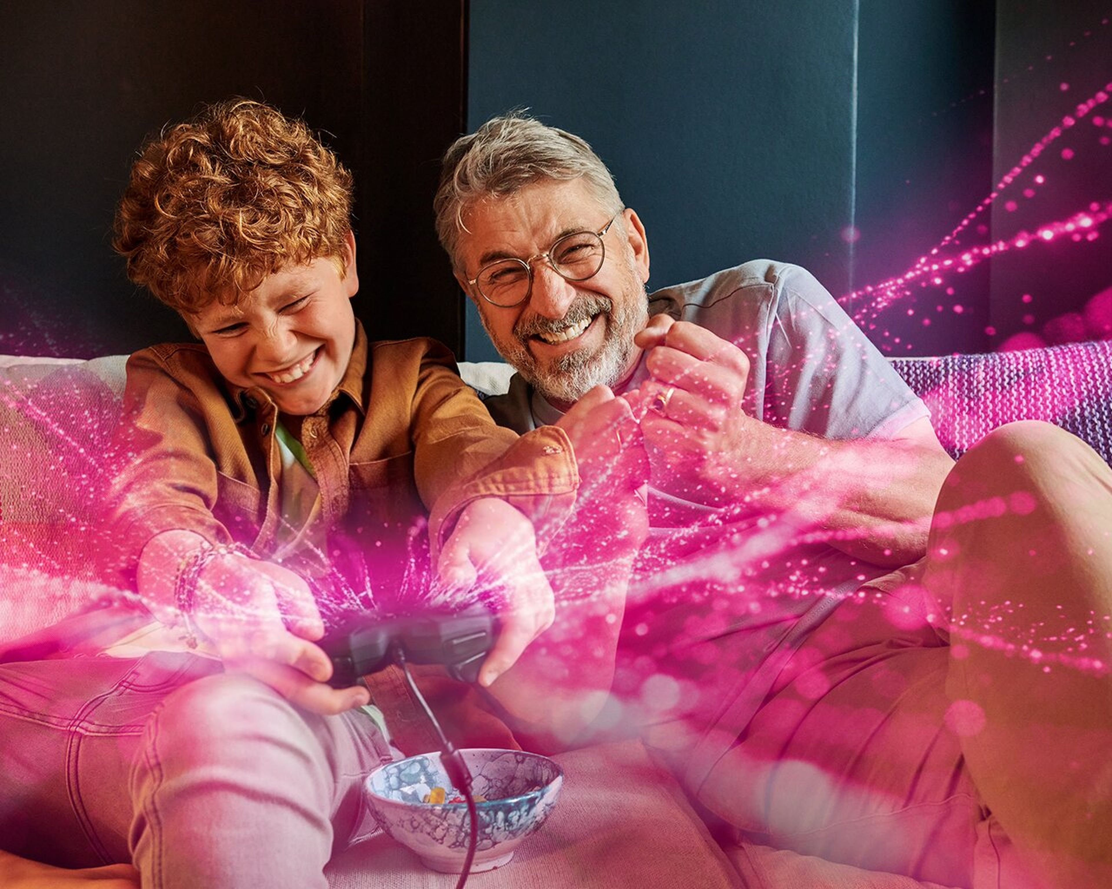 Radostné dieťa a starší muž hrajú spolu videohry na gauči, obklopení farebnými ružovými svetelnými efektmi.