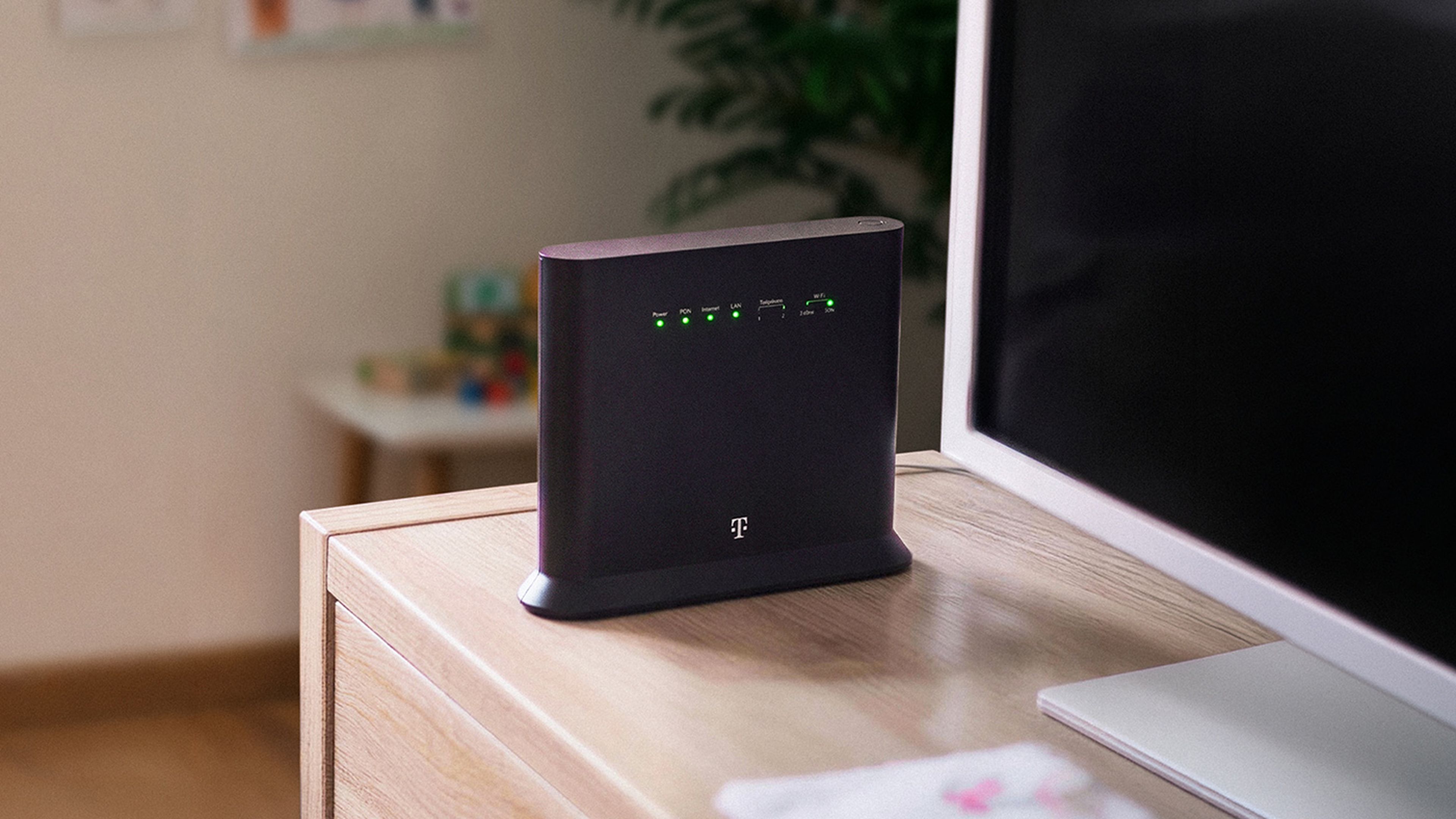Čierny router Telekomu na drevenej skrinke vedľa monitora.