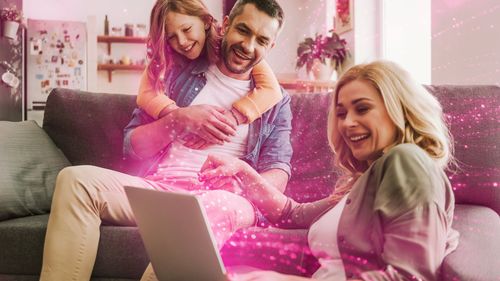 Trojčlenná rodina radostne sedí na gauči s otvoreným notebookom a ružové trblietky osvetľujú scénu a vytvárajú magickú atmosféru.