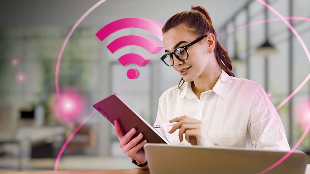 Žena v okuliaroch sedí pri notebooku a používa tablet a stylus. V pozadí je zobrazený ružový symbol Wi-Fi.