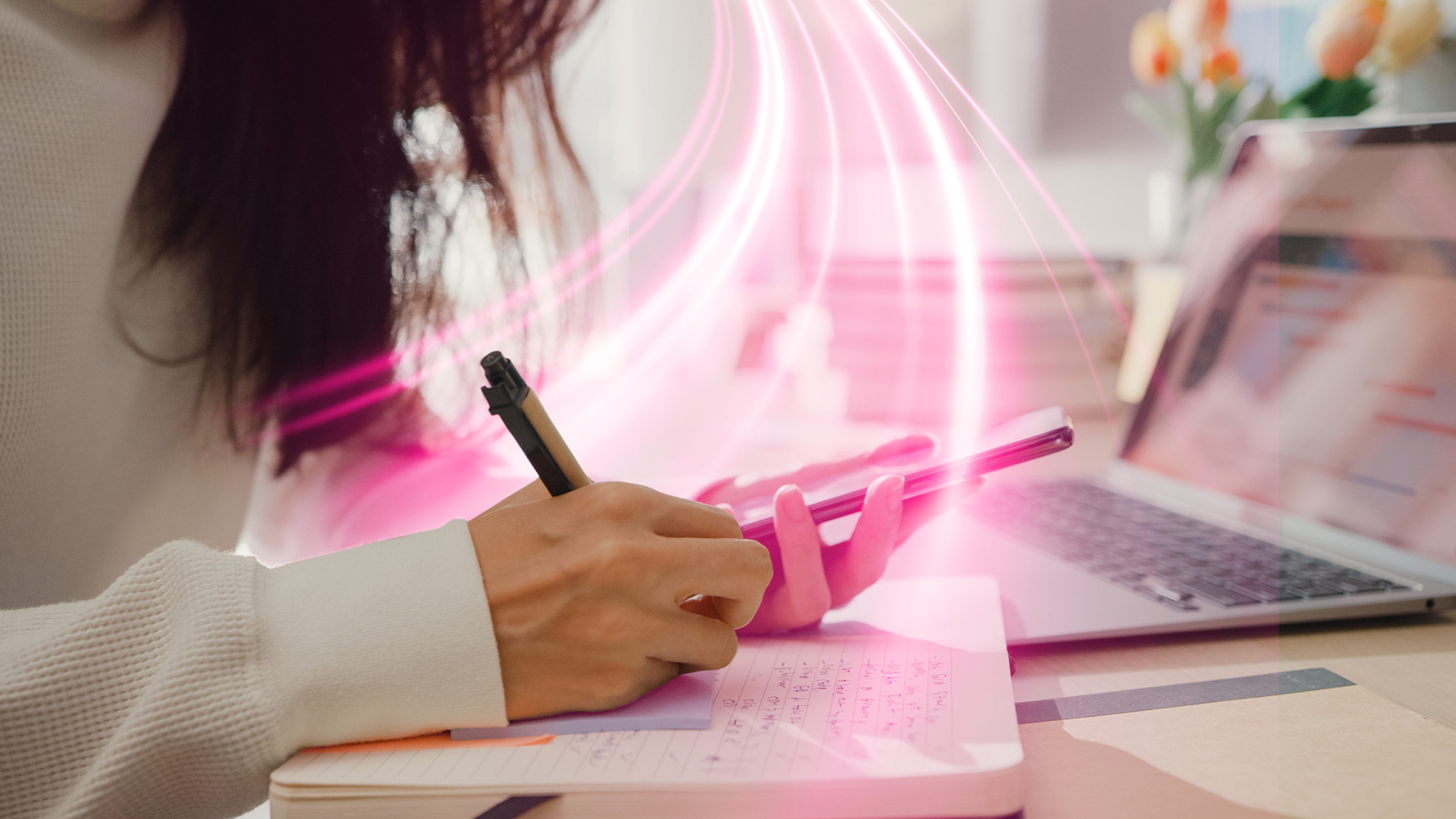 Žena píše do zápisníka a drží mobil, vedľa je notebook s ružovým svetlom.