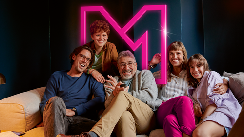 Skupina piatich ľudí usmieva na gauči pred ružovým logom "M".