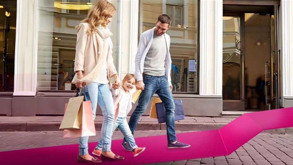 Trojčlenná rodina s nákupnými taškami v rukách sa šťastne prechádza po dláždených uliciach pred obchodom s veľkými oknami.