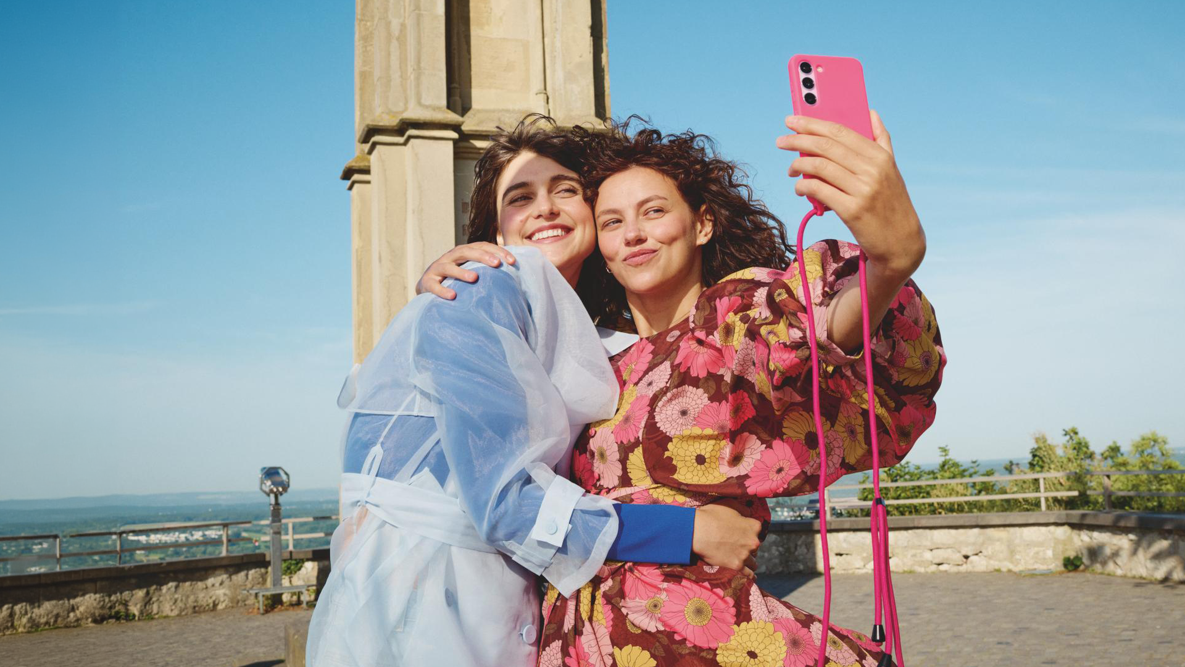 Dve ženy si robia selfie s ružovým telefónom, s úsmevom a objímajú sa, zatiaľ čo stoja pred historickou stavbou.