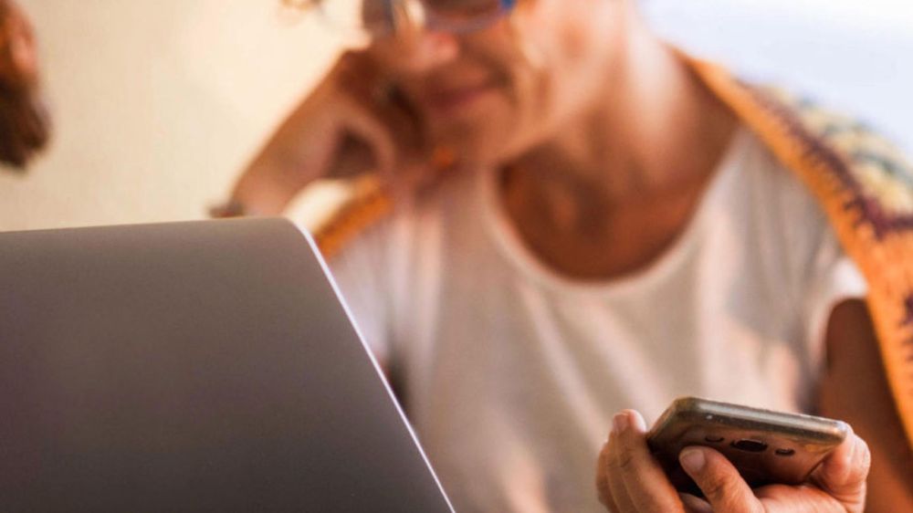 Žena v okuliaroch používa notebook a drží mobilný telefón, prikrytá farebnou háčkovanou dekou.