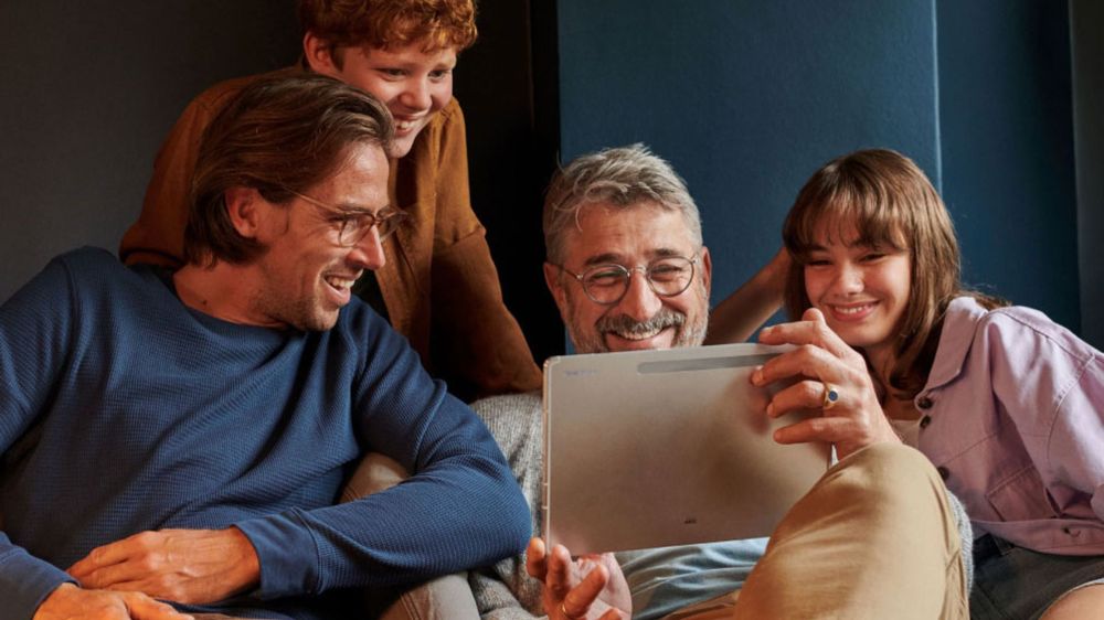 Štvorčlenná rodina, vrátane dvoch dospelých a dvoch detí, sa usmieva a pozerá na tablet, zatiaľ čo sedí na pohovke v útulnej izbe.
