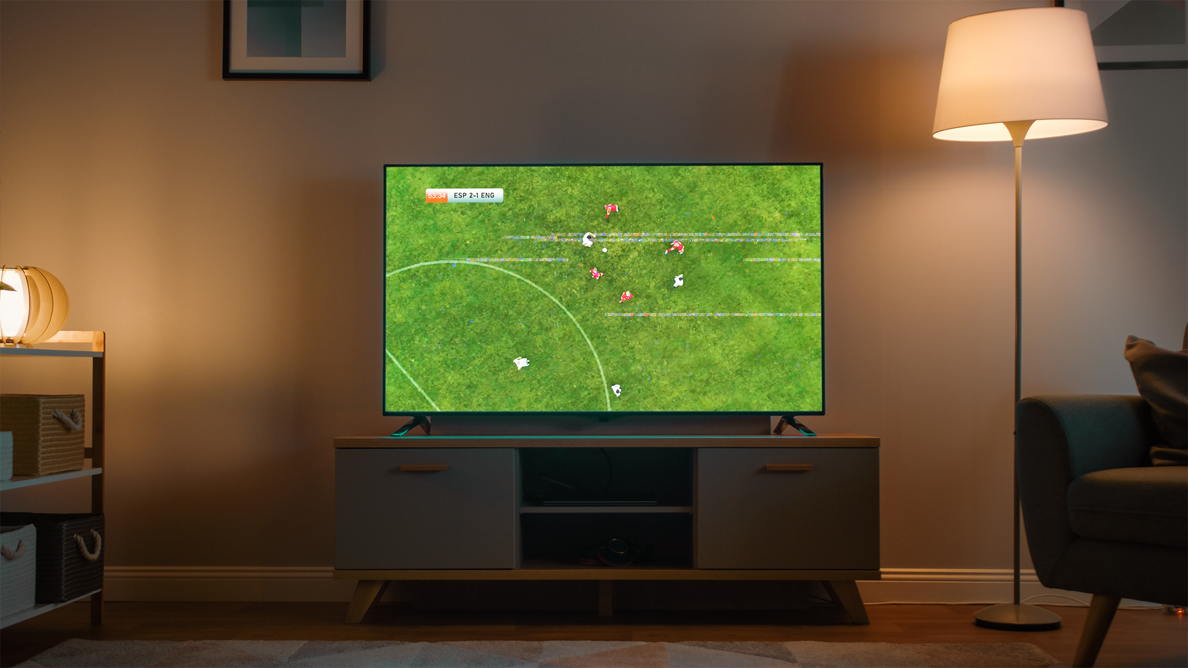 Televízor v obývačke zobrazuje futbalový zápas, okolo je lampička a polica s košíkmi.