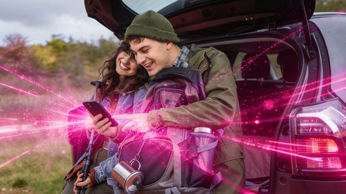 Dvaja ľudia v outdoorovom oblečení sa opierajú o otvorený kufor auta, smejú sa a pozerajú sa na telefón. Z tohto zariadenia vychádzajú ružové energetické lúče.
