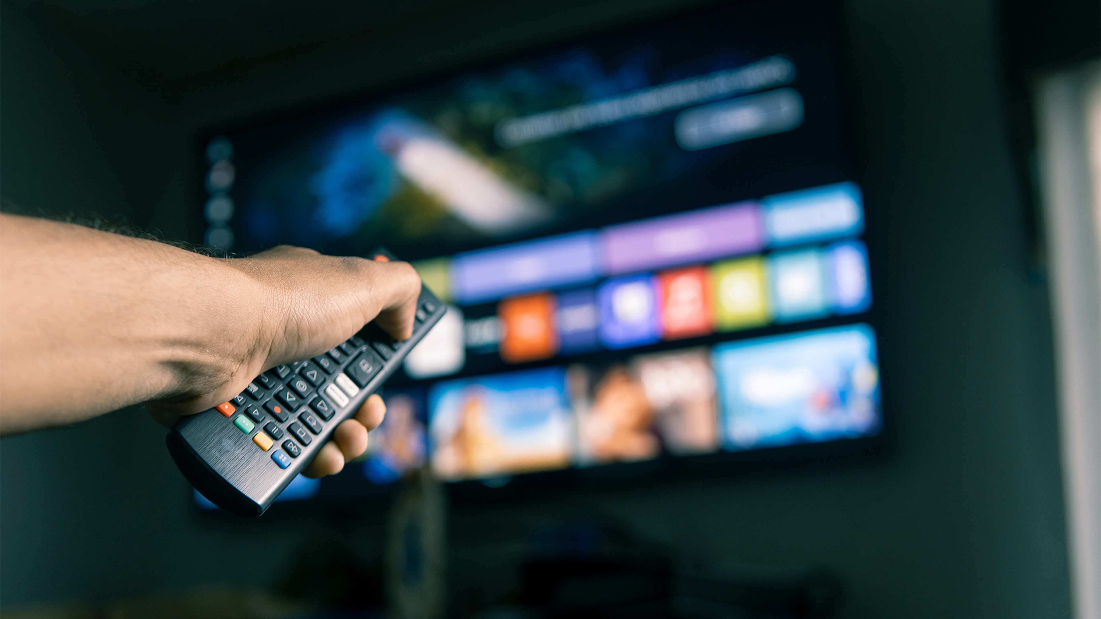 Ruka držiaca diaľkový ovládač pred televíznou obrazovkou zobrazujúcou menu aplikácií.