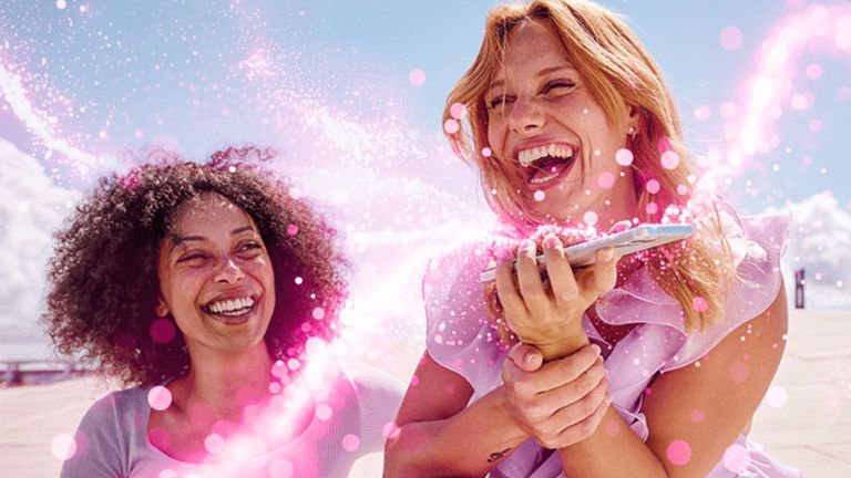 Dve ženy sedia vonku, usmievajú sa a smejú sa, obklopené ružovými, žiariacimi vírmi a trblietkami, s jasnou oblohou v pozadí.