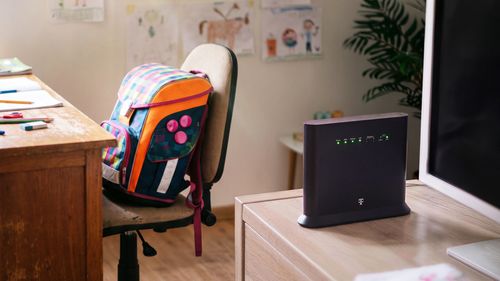 Školská taška na stoličke vedľa stola s routerom Telekom a počítačom.