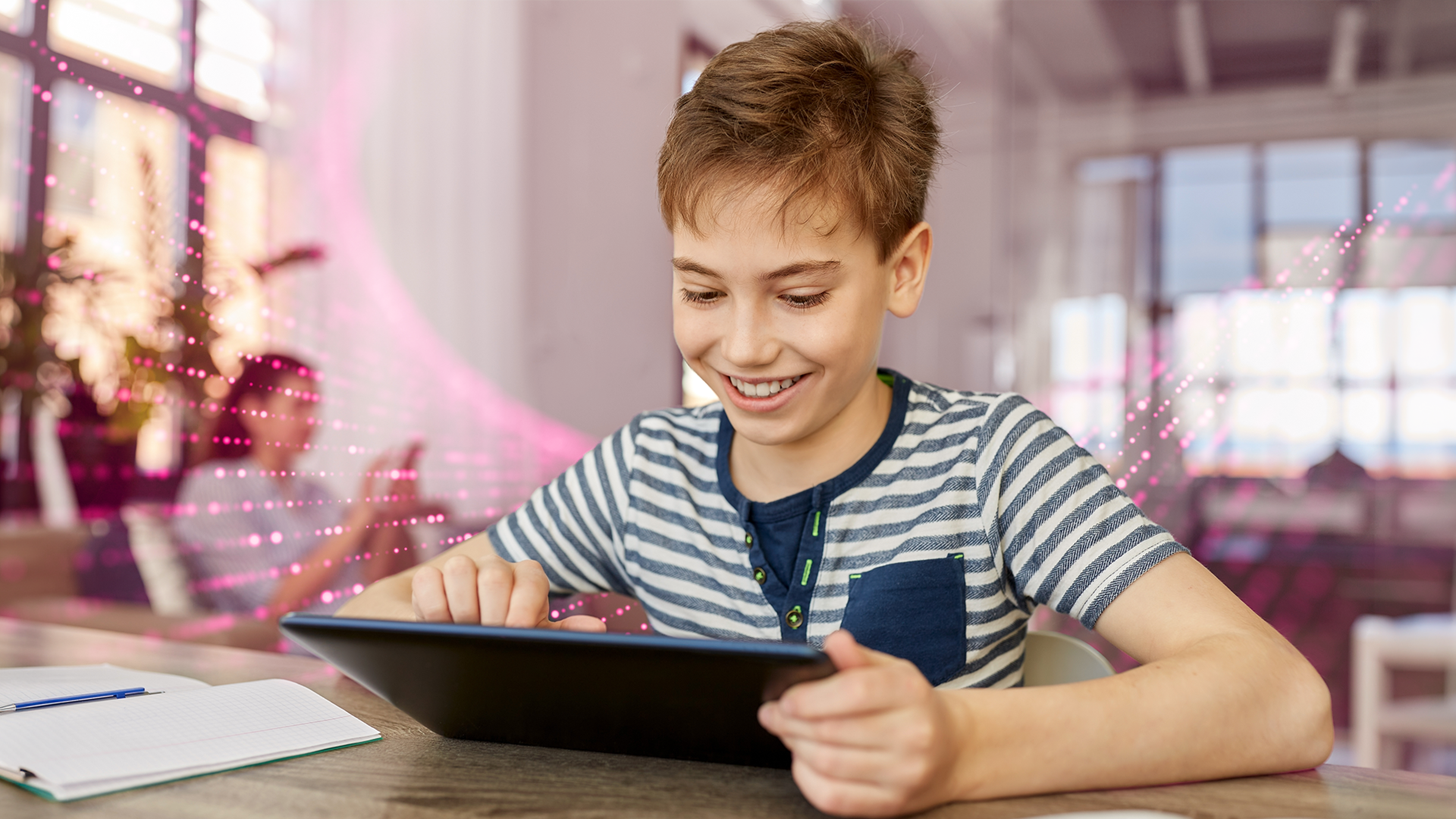 Chlapec so šťastným výrazom pracuje na tablete, sedí pri stole s poznámkovým blokom a perom, okolo digitálne efekty.