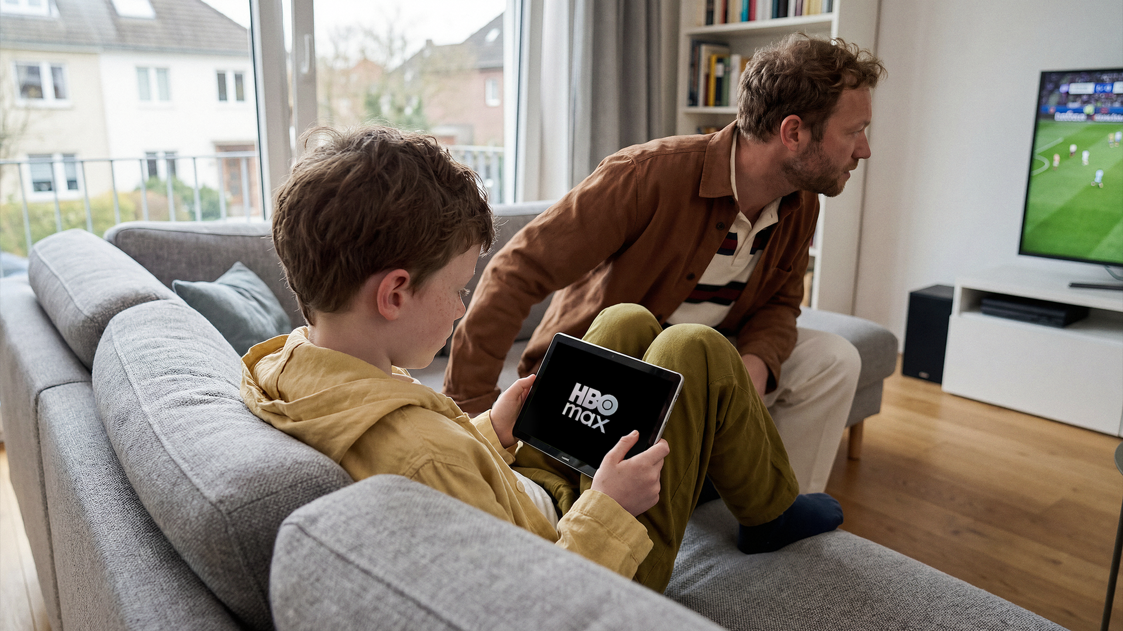 Chlapec sleduje HBO Max na tablete a muž sedí vedľa neho, pričom sleduje futbalový zápas na televízore.