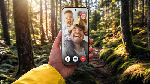 Ruka držiaca mobil s videohovorom v lese, usmievajúci sa muž a dieťa na displeji.