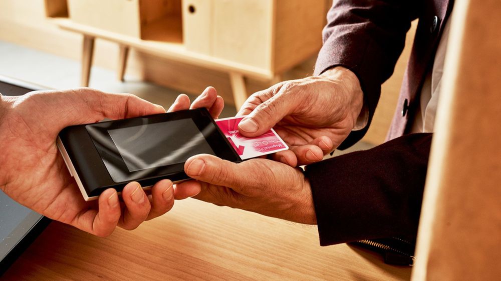 Osoba podáva malú kartu inej osobe držiacej smartfón, pravdepodobne na digitálnu transakciu, pri drevenom pulte.