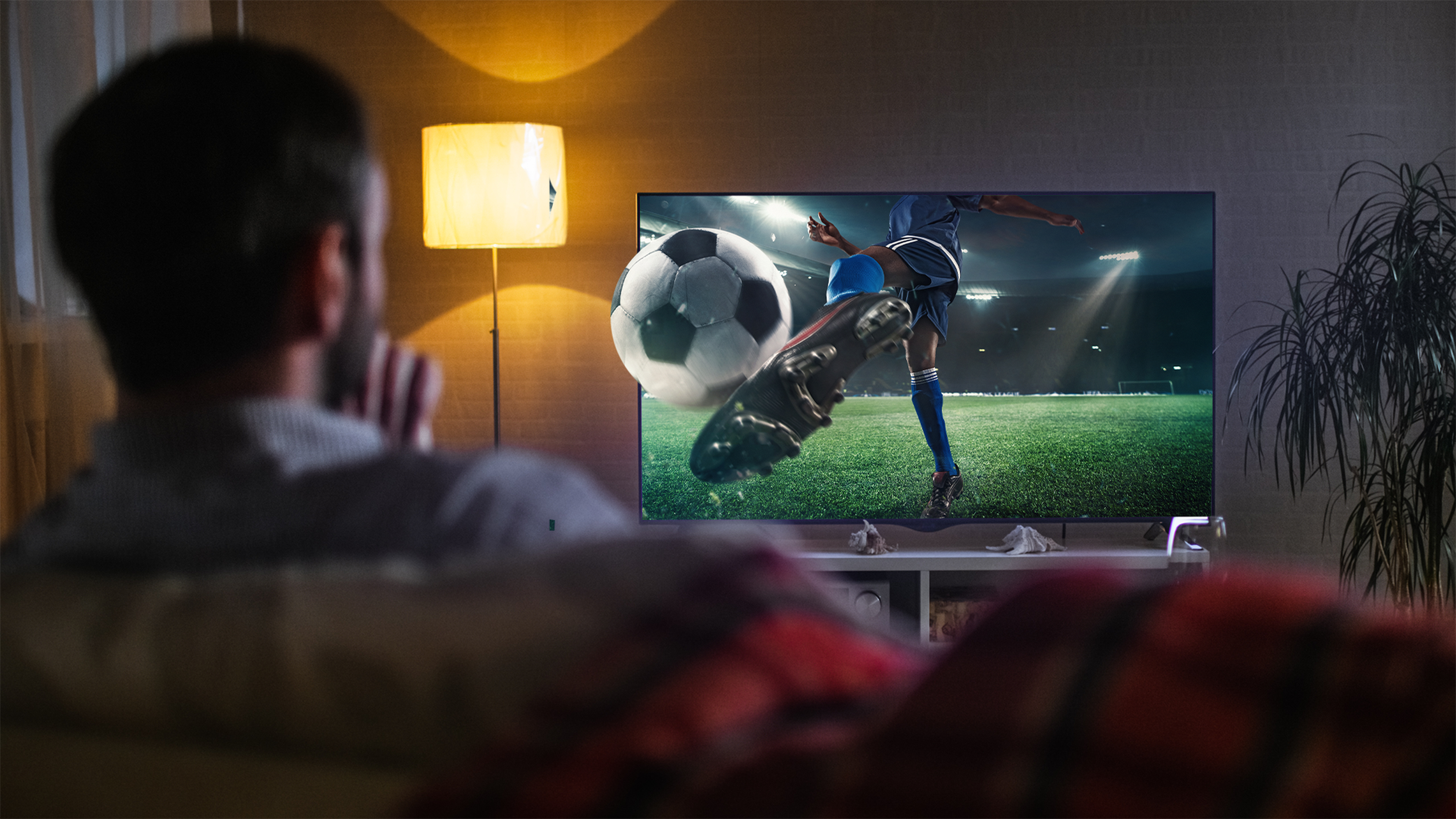 Muž sleduje futbalový zápas v televízii, kde hráč kopne loptu.