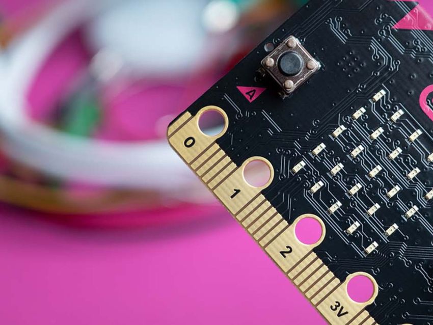 Detailný záber dosky mikrokontroléra s tlačidlami a zlatými konektormi na rozmazanom ružovom pozadí s drôtmi.