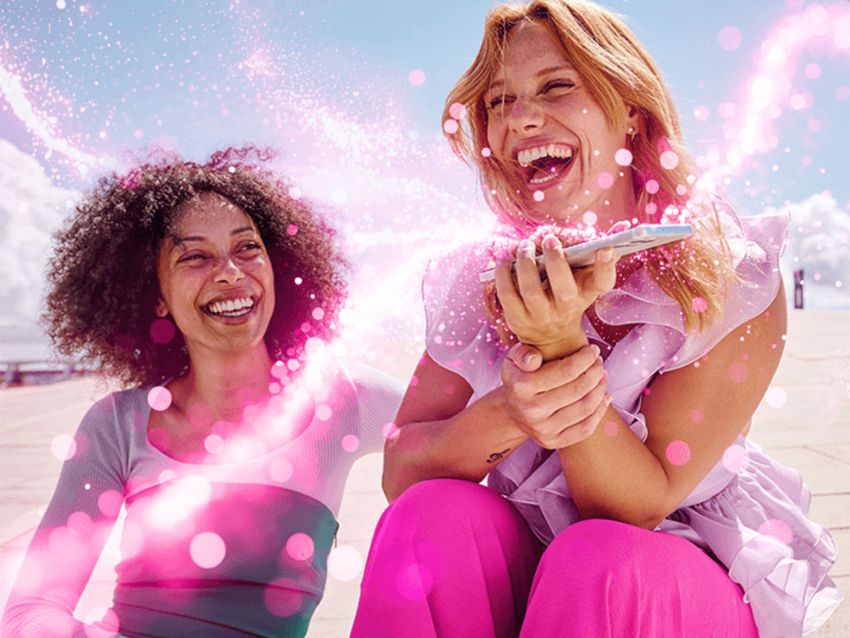 Dve ženy sedia vonku, usmievajú sa a smejú sa, obklopené ružovými, žiariacimi vírmi a trblietkami, s jasnou oblohou v pozadí.