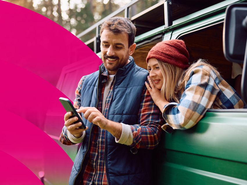 Usmievavý pár sa spolu pozerá na smartfón, vykláňajúc sa zo zeleného vozidla v zalesnenej oblasti. Žena má na sebe červenú čiapku a kockovanú košeľu.