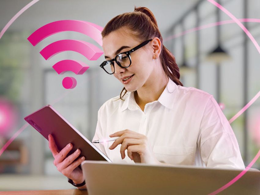 Žena v okuliaroch sedí pri notebooku a používa tablet a stylus. V pozadí je zobrazený ružový symbol Wi-Fi.
