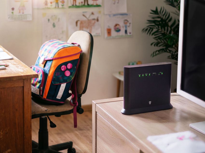 Router na stole vedľa počítača, opodiaľ farebný ruksak na stoličke.