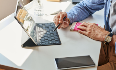 Muž pracuje s tabletom a klávesnicou na stole, drží pero v jednej ruke a knihu v druhej. Na stole leží smartfón.