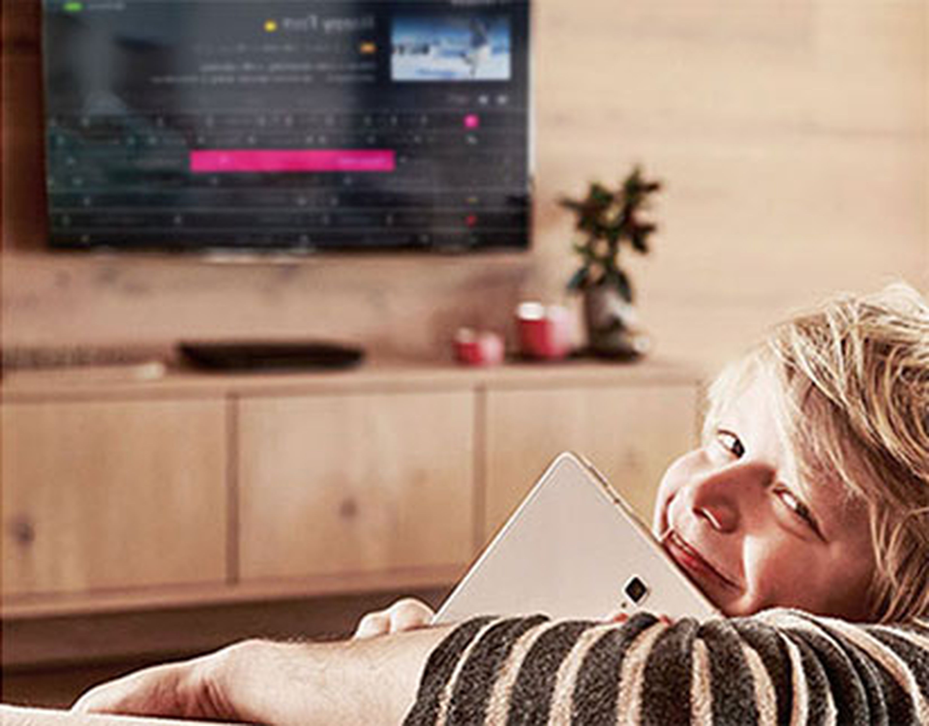 Usmievavá osoba sediaca na gauči s tabletom v ruke. V pozadí na drevenej stene je rozmazaná televízna obrazovka.