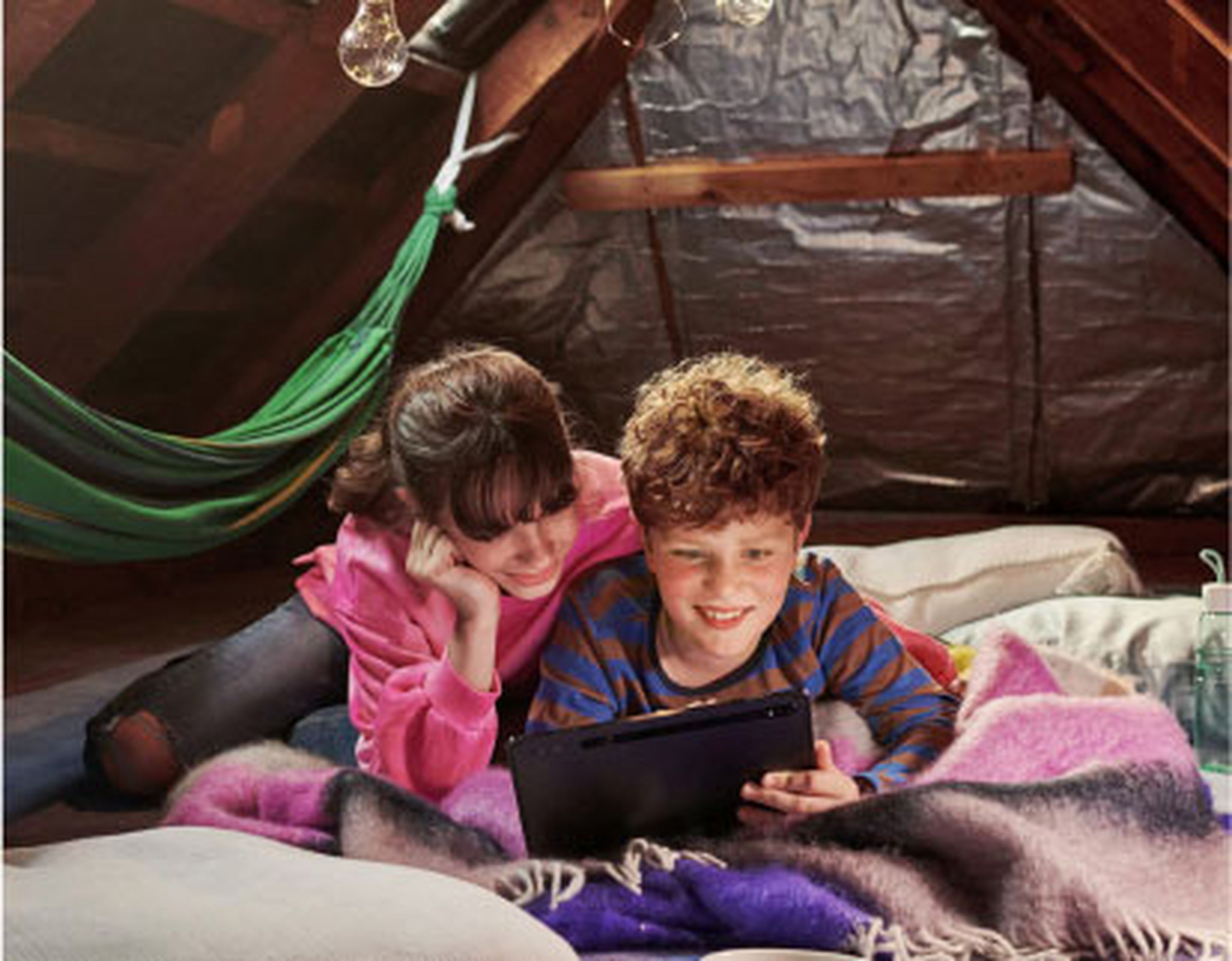 Dve deti ležia na deke na povale, usmievajú sa a pozerajú sa na tablet, v pozadí je hojdacia sieť a visiace lampy.