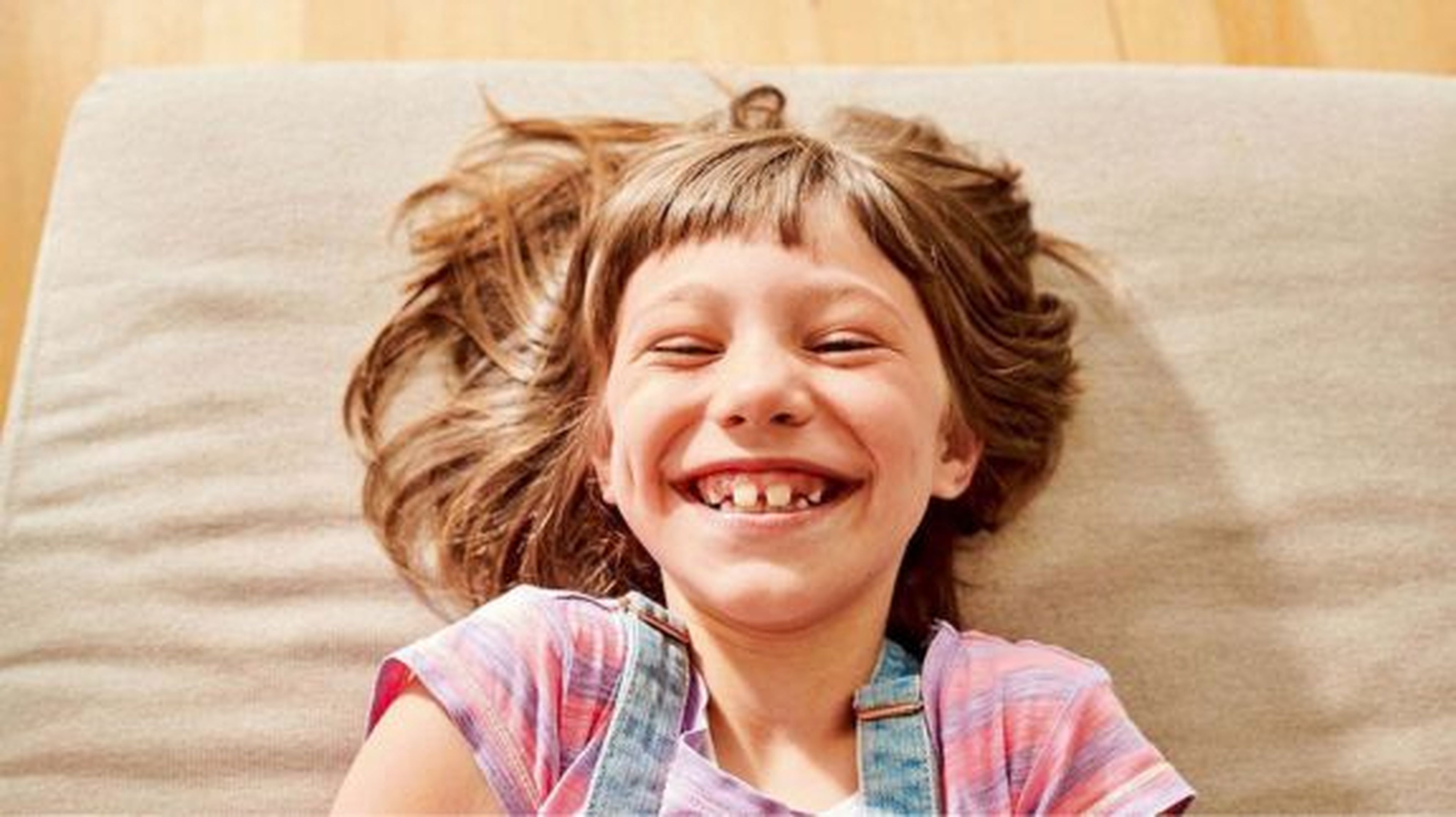 Usmievavé dieťa s hnedými vlasmi leží na svetlom vankúši, má na sebe ružové tričko a džínsový overal.