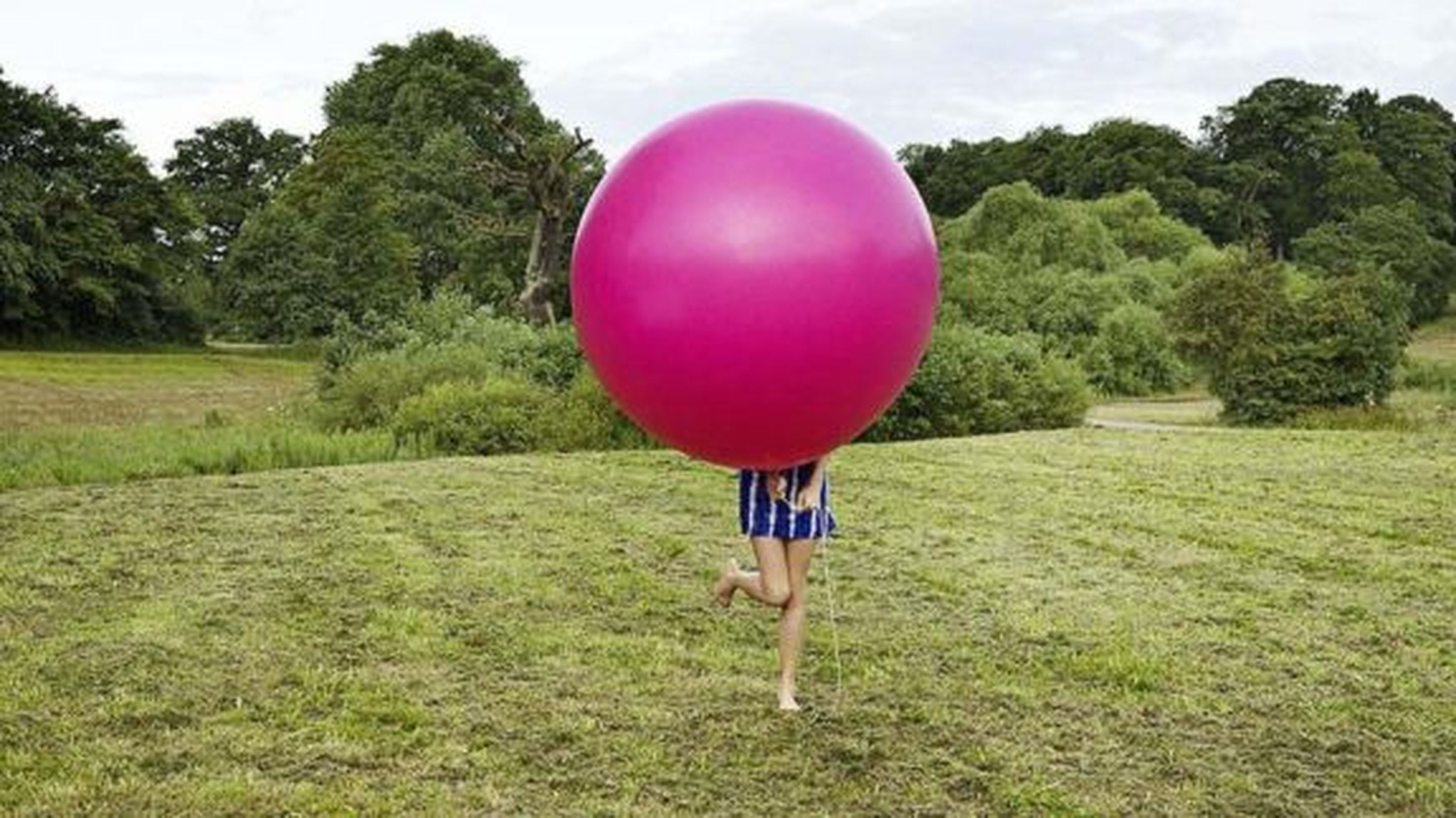 Osoba bežiaca po trávnatom poli, väčšinou skrytá za veľkou ružovou loptou, so stromami v pozadí.