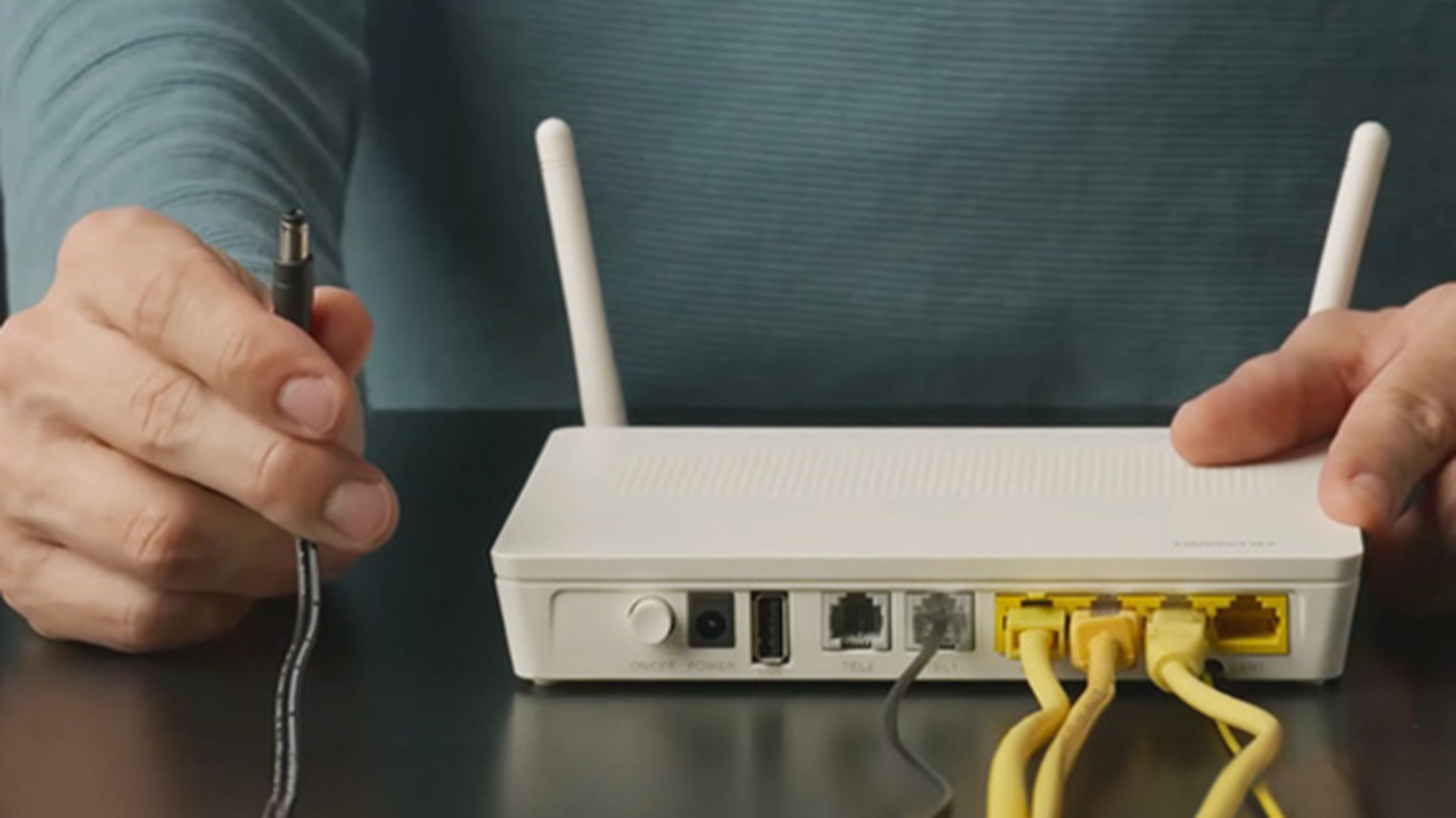Osoba pripája kábel k bielemu routeru s dvoma anténami, pričom je doň zapojených viacero žltých ethernetových káblov.