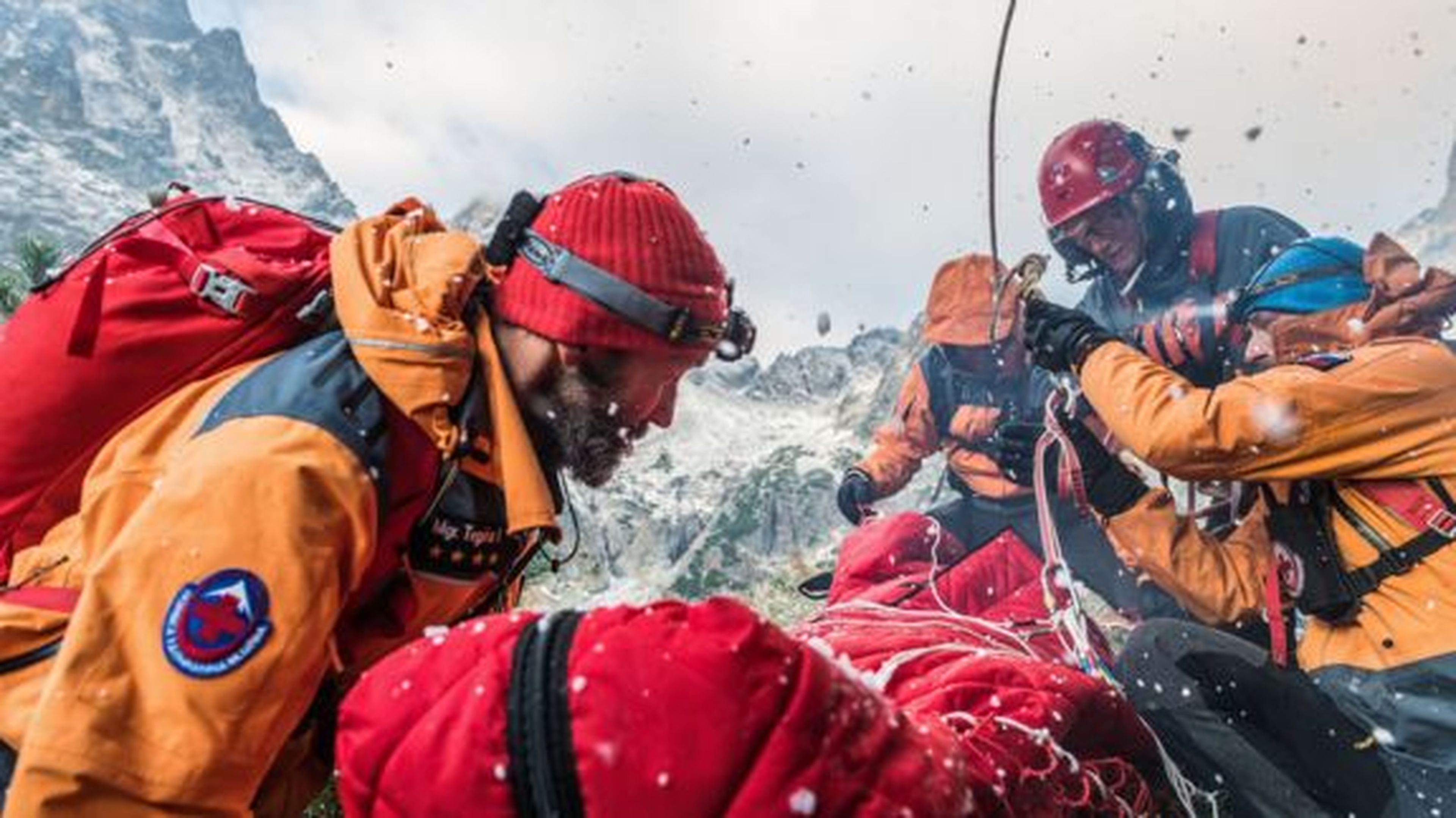 Záchranný tím v oranžových bundách a červených prilbách zaisťuje nosidlá lanami v zasneženom horskom teréne. Okolo nich padajú snehové vločky.