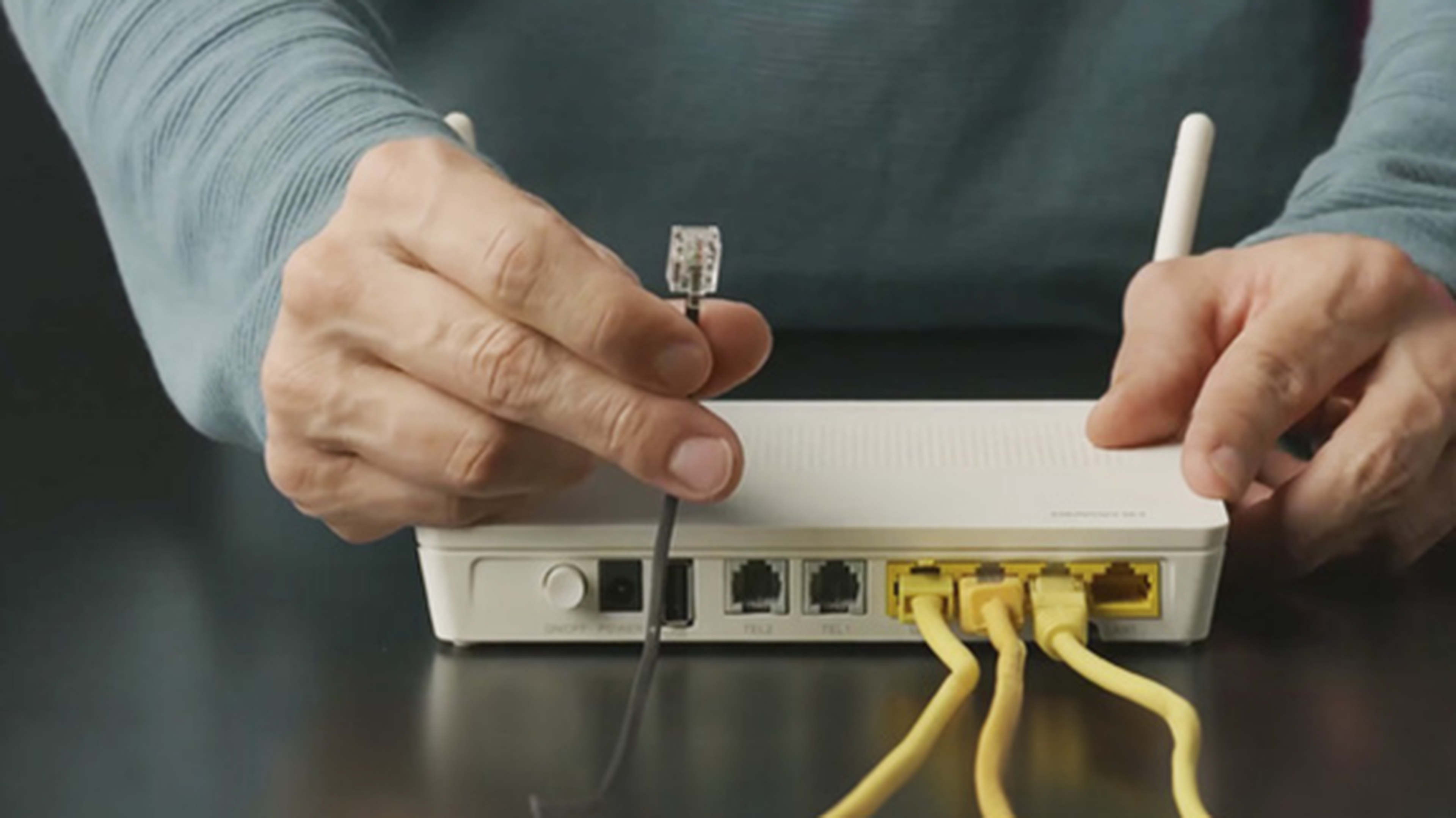 Osoba pripájajúca ethernetový kábel k bielemu routeru s viacerými portami a žltými káblami na tmavom povrchu.